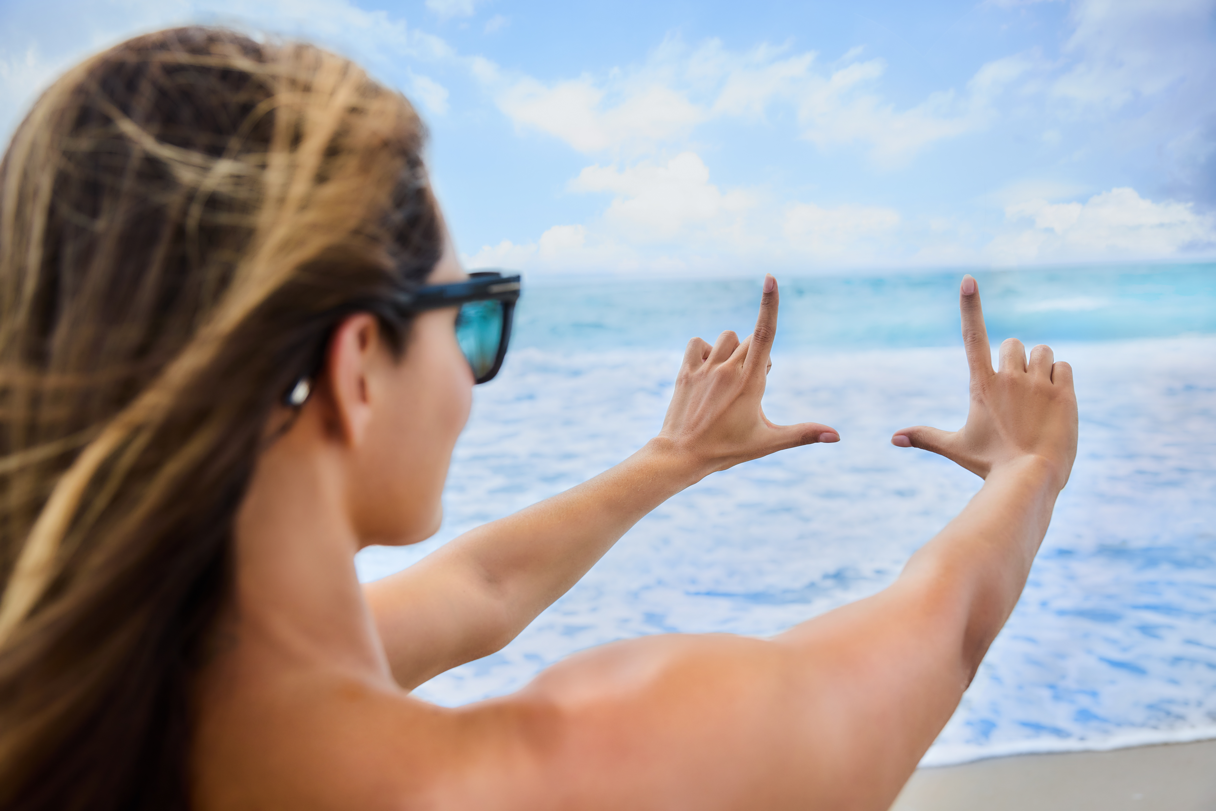 Woman in sunglasses framing ocean view with hands against blue sky and sea background.