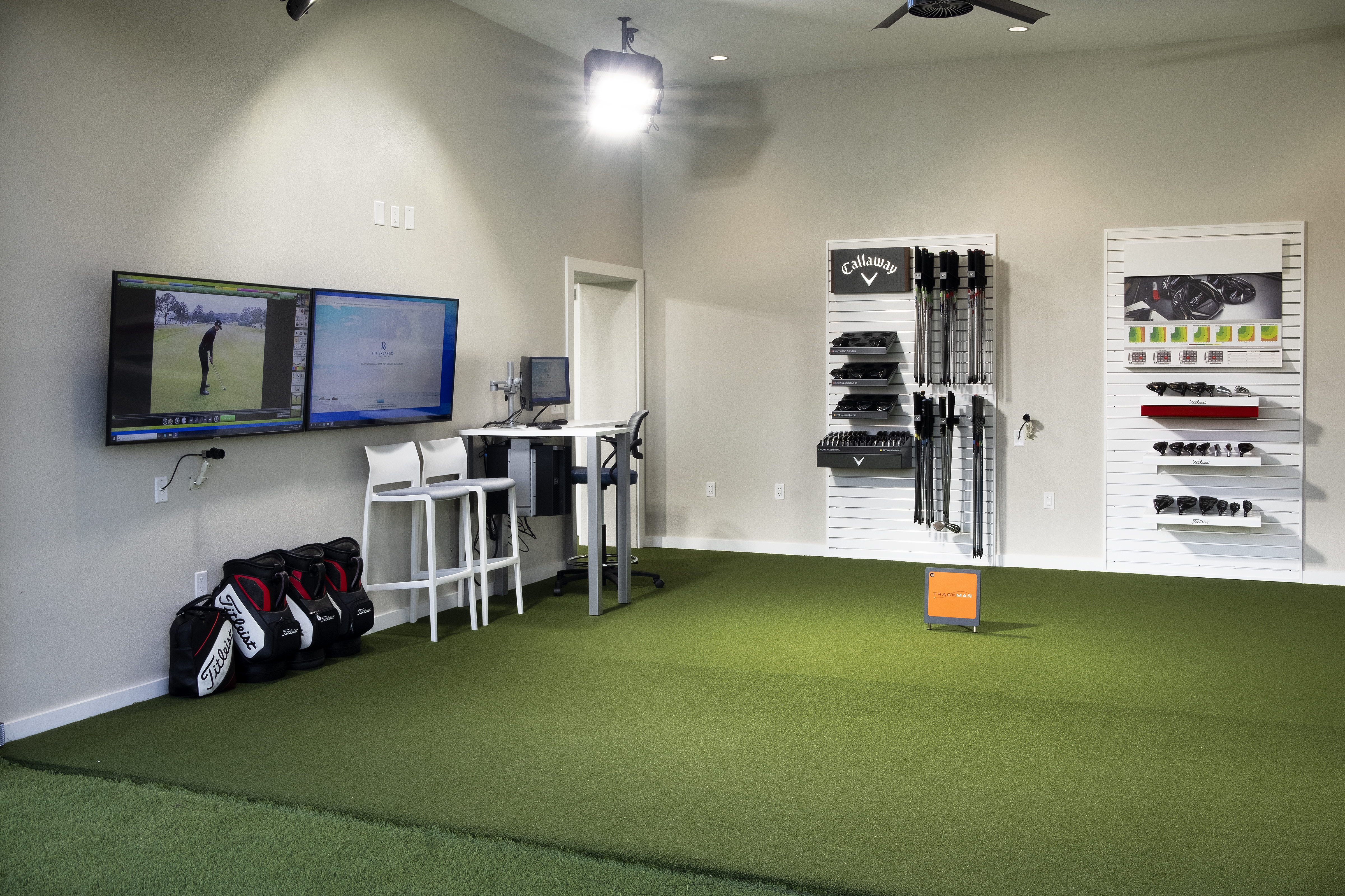 Indoor golf simulator room with green turf, dual monitors, golf equipment on walls, and golf bags.