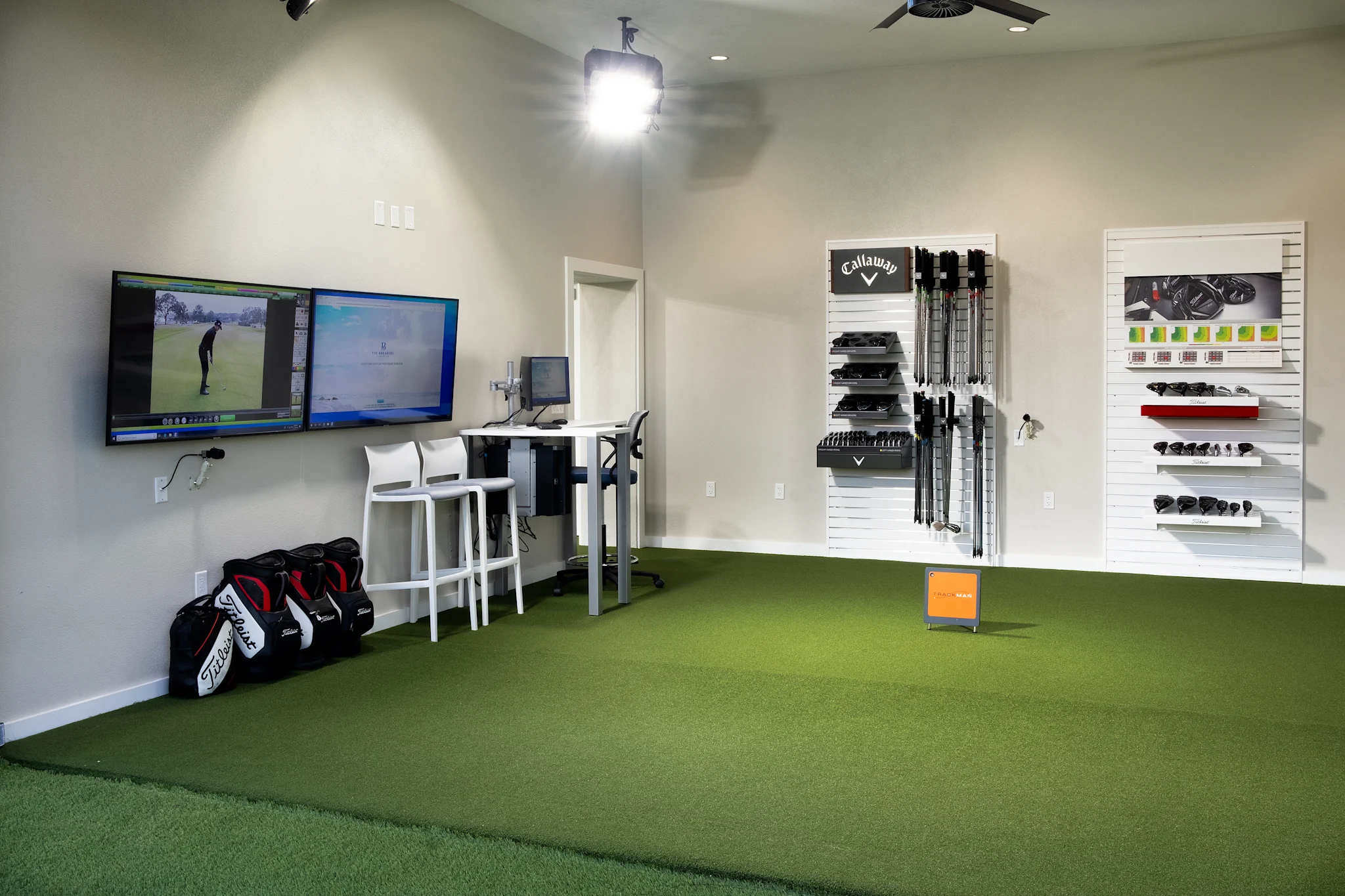 Indoor golf simulator room with green turf, dual monitors, golf equipment on walls, and golf bags.
