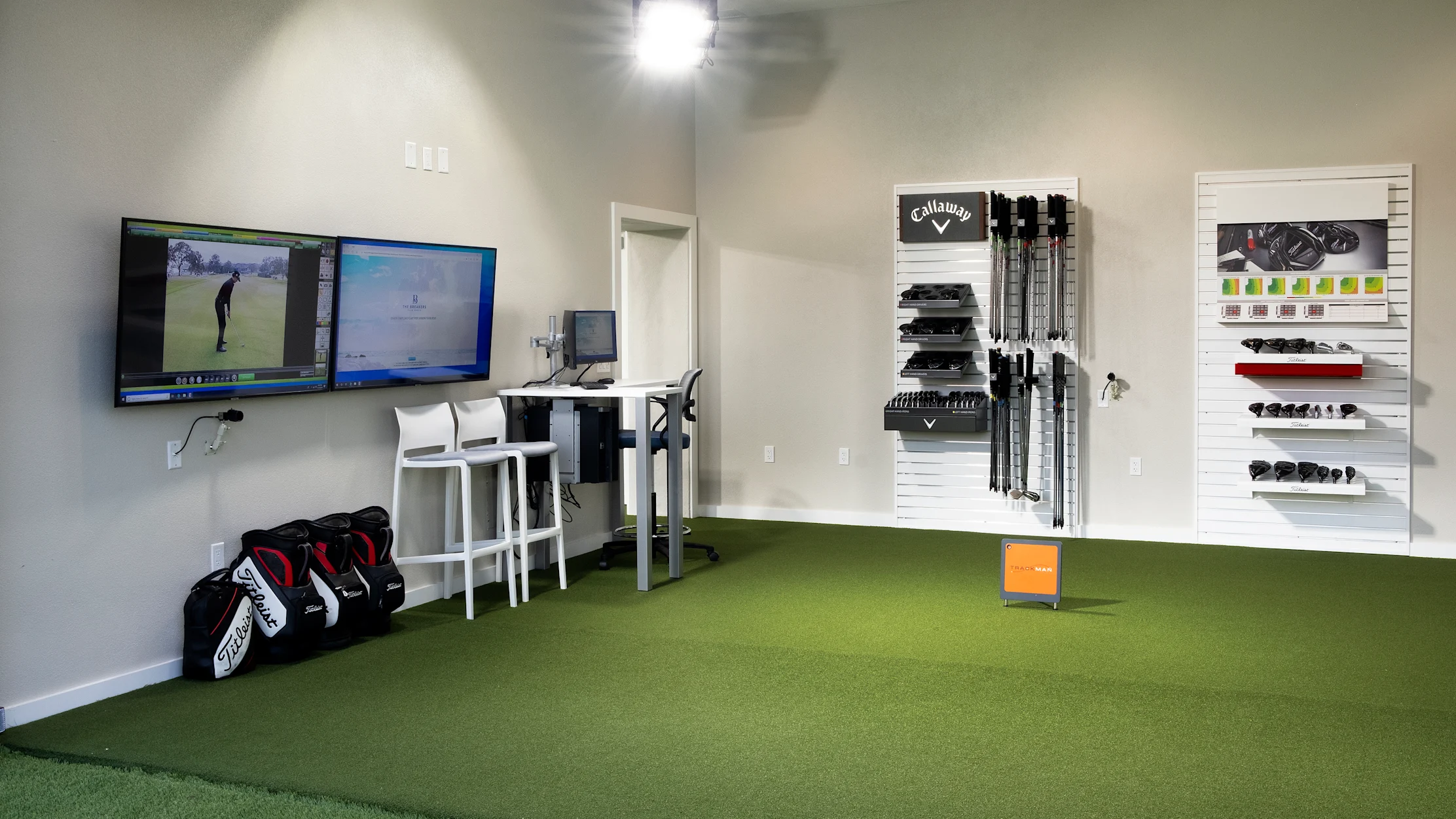 Indoor golf simulator room with green turf, dual monitors, golf equipment on walls, and golf bags.