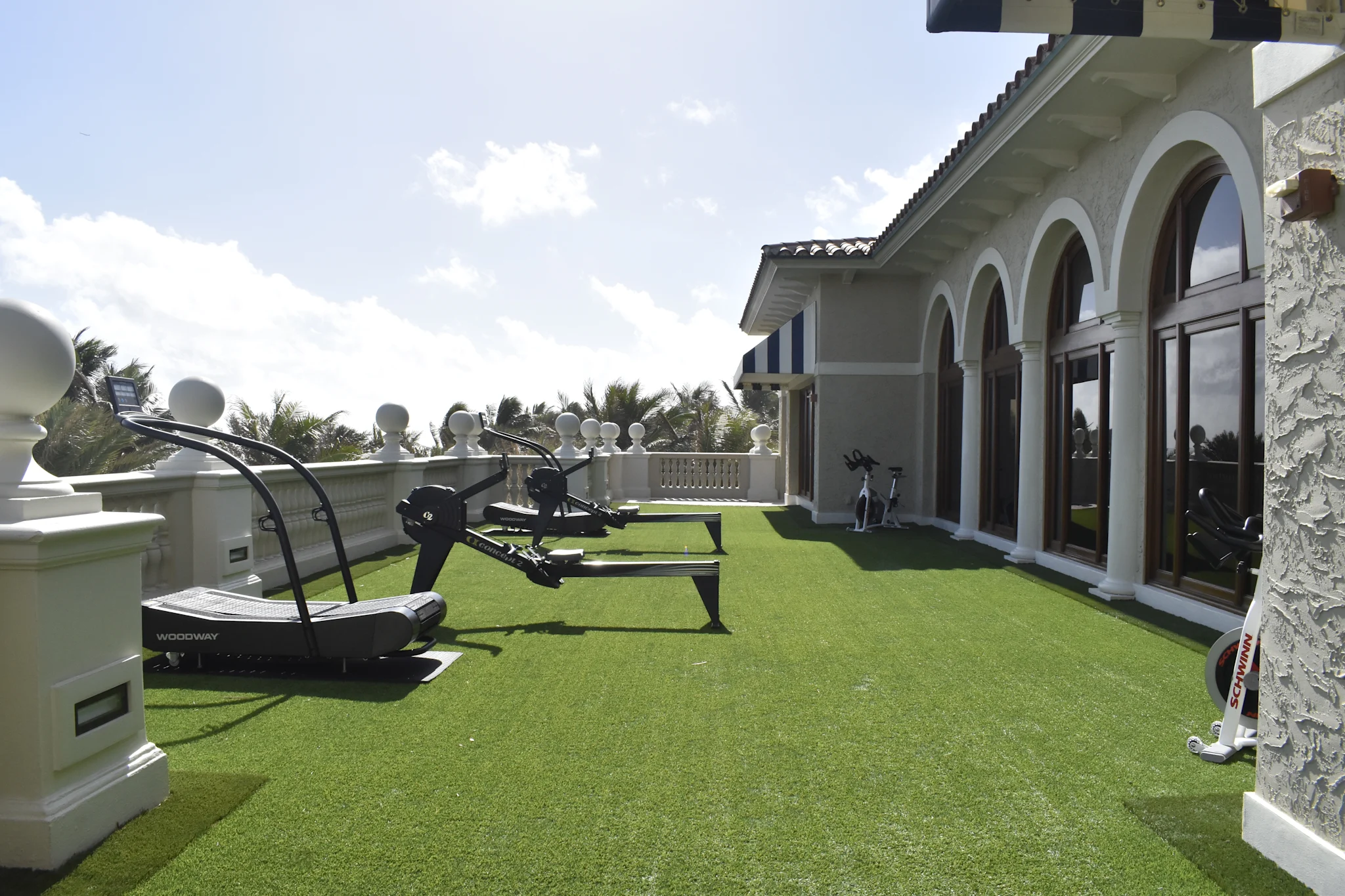 Rooftop terrace with green artificial turf, exercise equipment, white arched building, and palm trees.