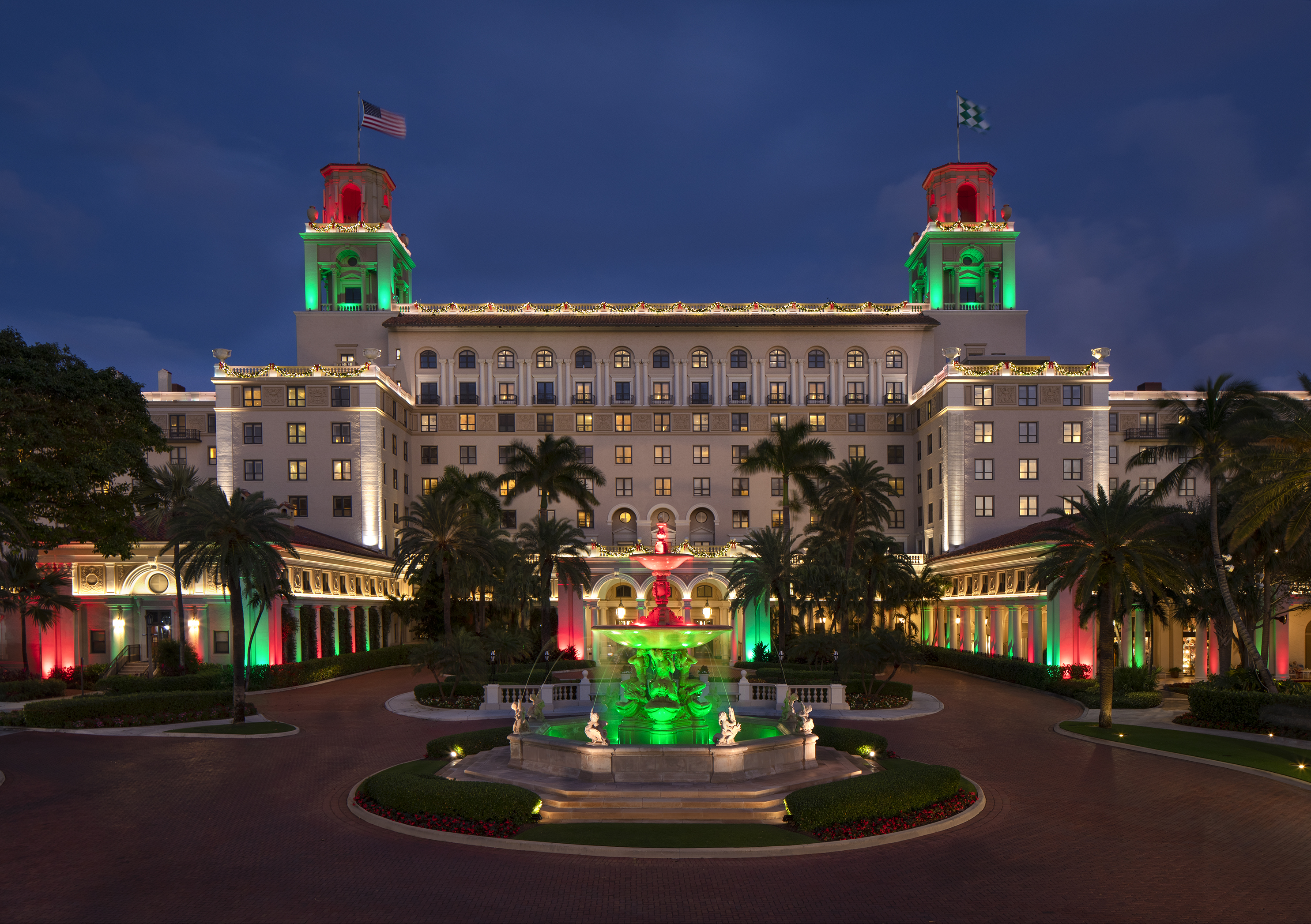 Grand hotel illuminated with red and green lights at dusk, featuring twin towers, fountain, and palm trees.