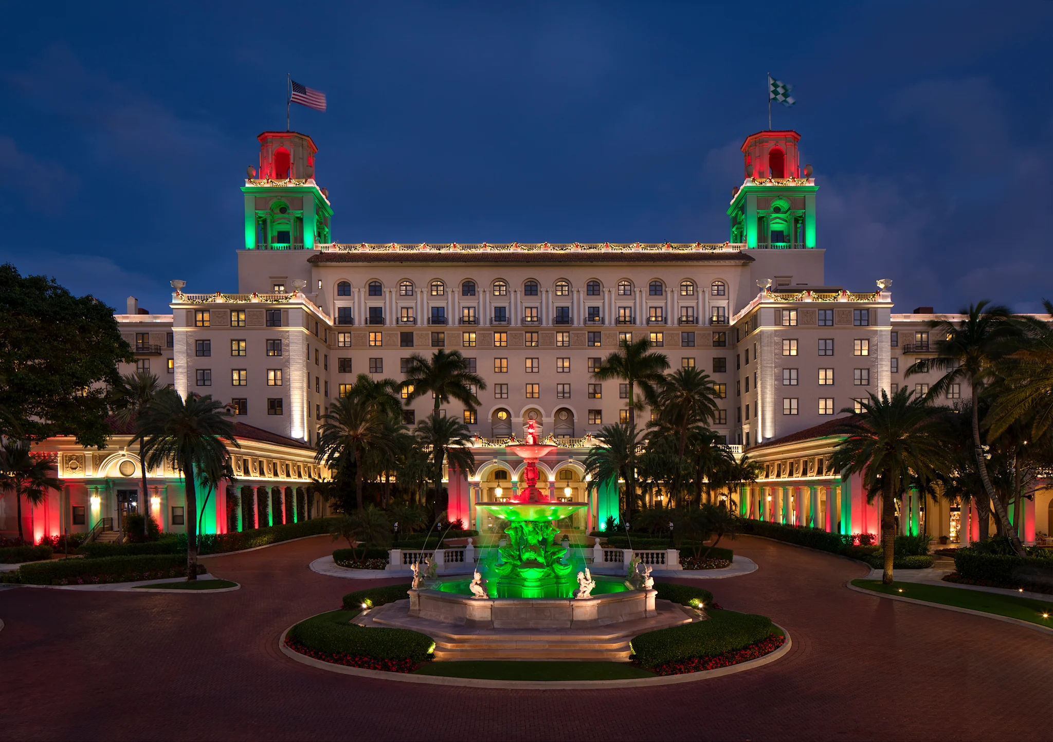 Grand hotel illuminated with red and green lights at dusk, featuring twin towers, fountain, and palm trees.