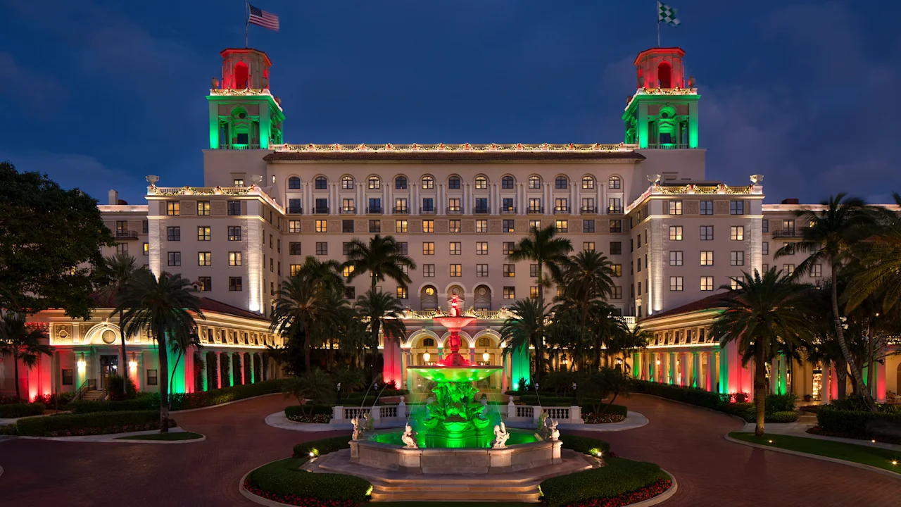 Grand hotel illuminated with red and green lights at dusk, featuring twin towers, fountain, and palm trees.