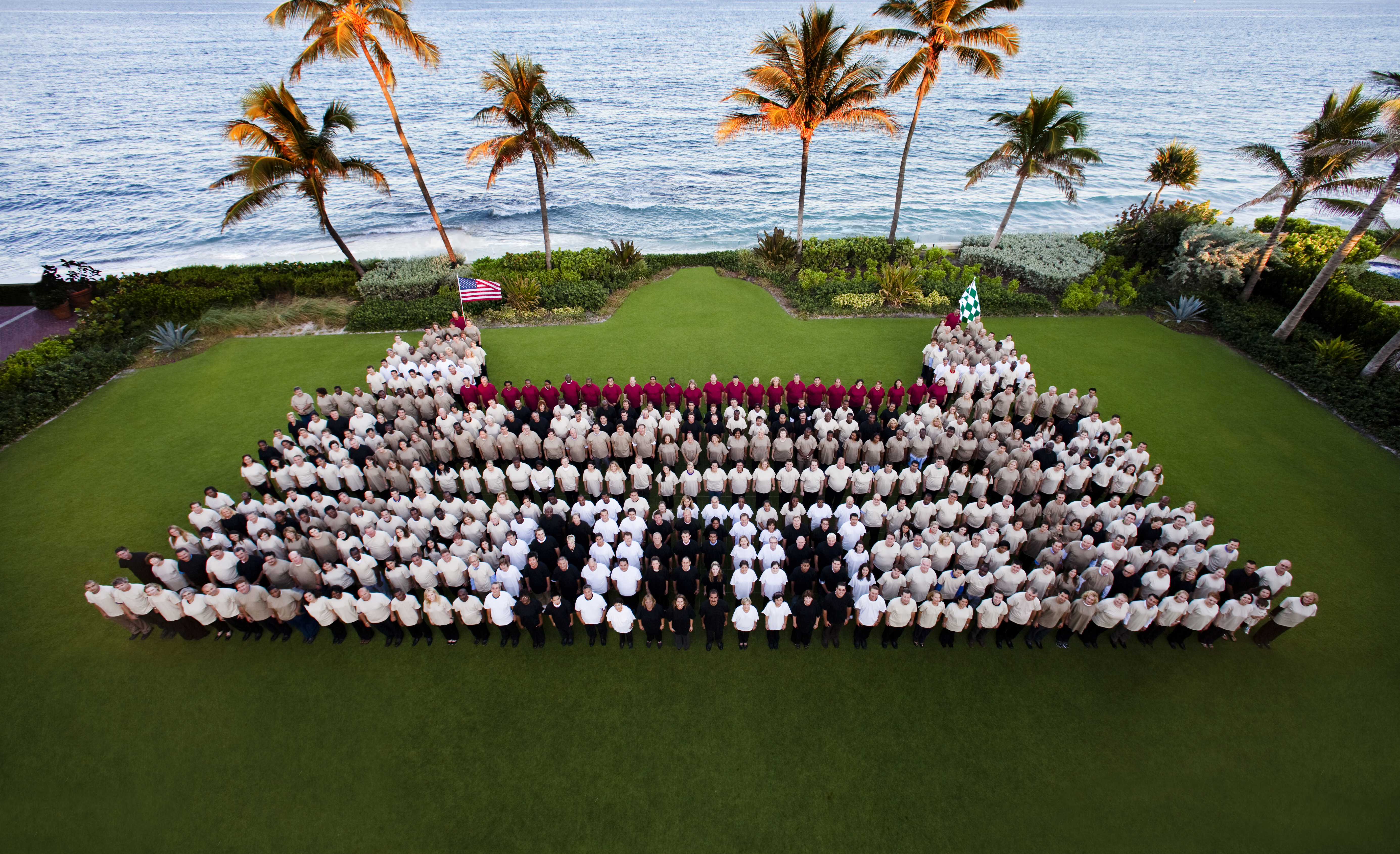 Large group photo of staff in uniform arranged on oceanfront lawn with palm trees and blue water.