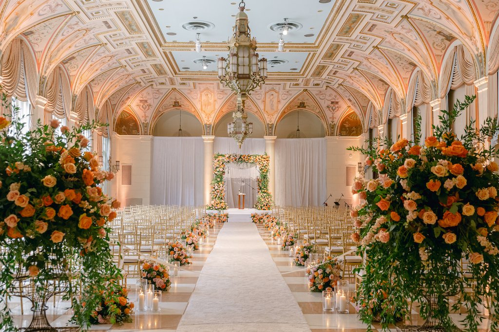 Elegant wedding ceremony venue with ornate ceiling, white aisle, gold chairs, and orange floral arrangements.