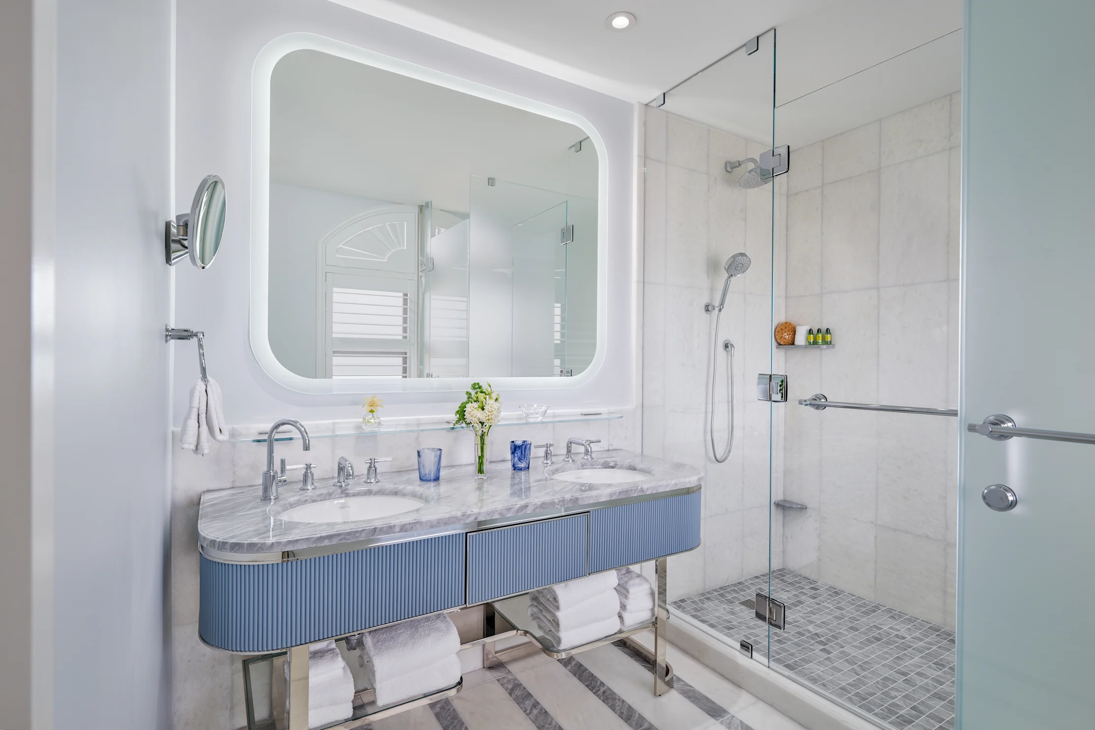 Modern bathroom with backlit mirror, blue curved vanity, marble countertop, and glass-enclosed shower.