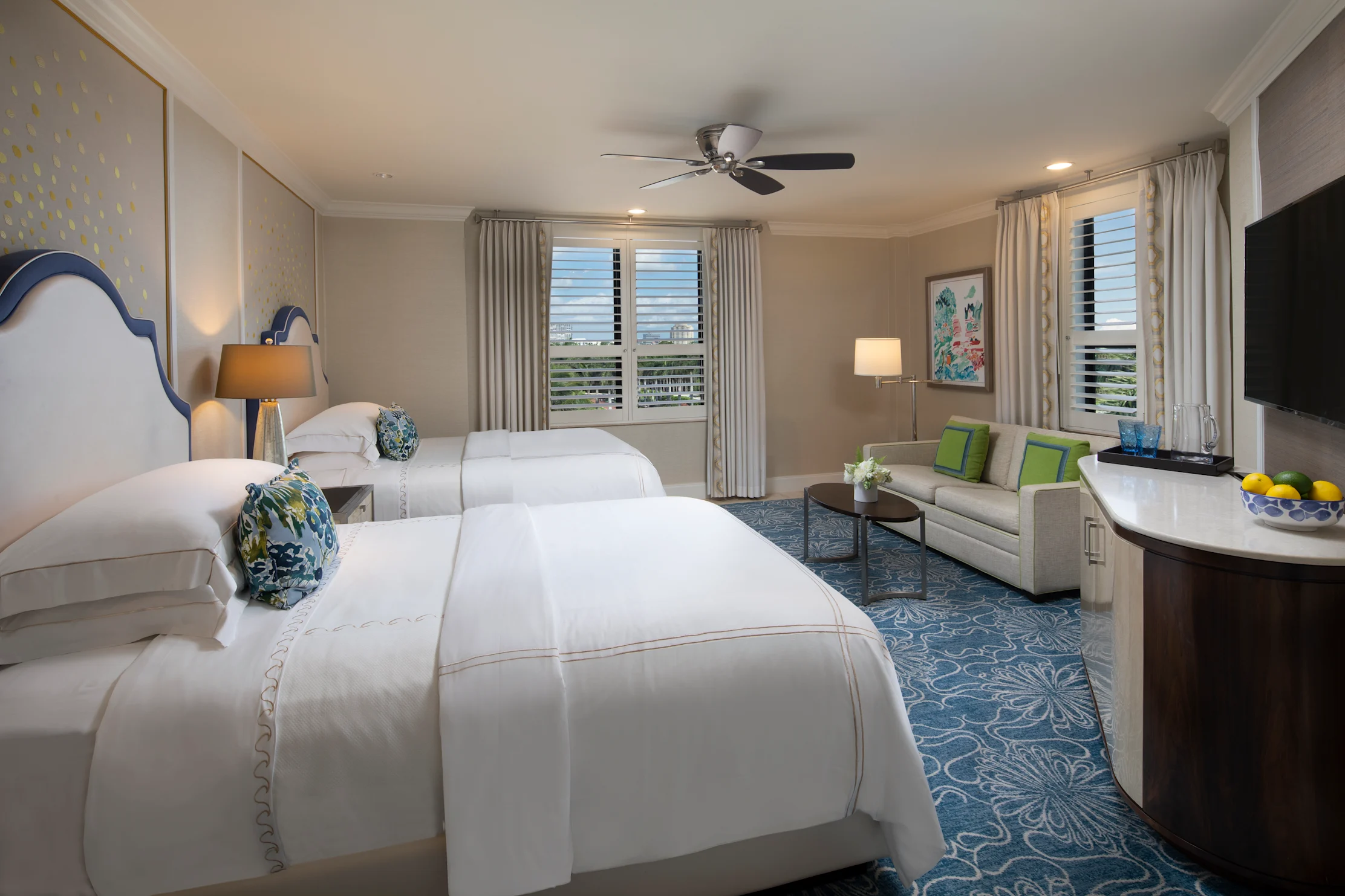 Elegant hotel room with two white beds, blue accents, seating area, and shuttered windows.