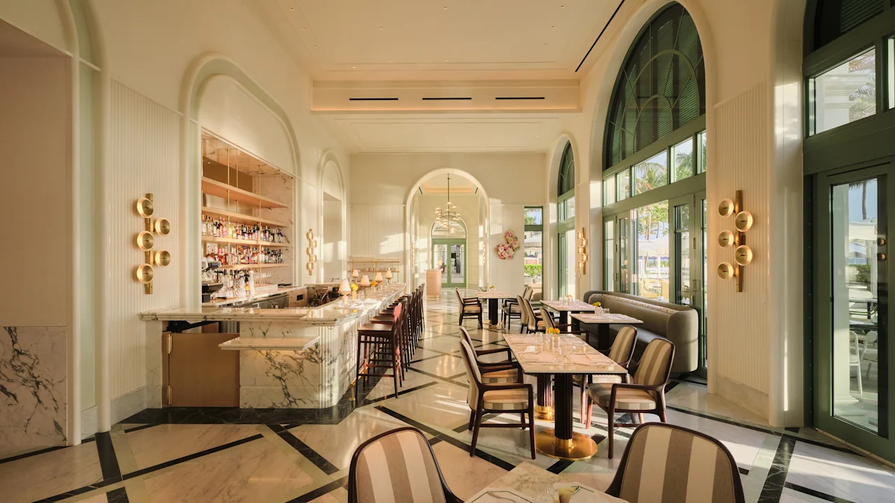 The sunlit Beach Club dining room, with tall arched windows, a marble bar and striped chairs