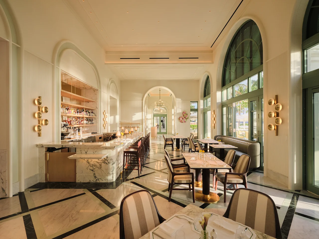 The sunlit Beach Club dining room, with tall arched windows, a marble bar and striped chairs