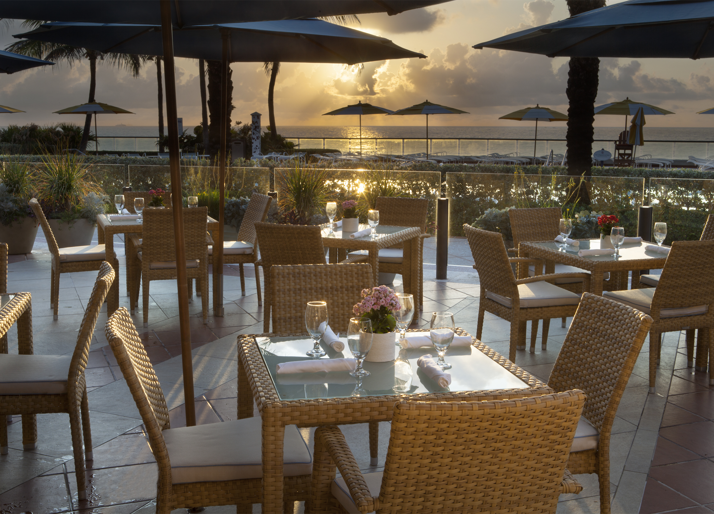 Oceanfront restaurant patio with wicker dining tables at sunset, blue umbrellas and palm trees visible.