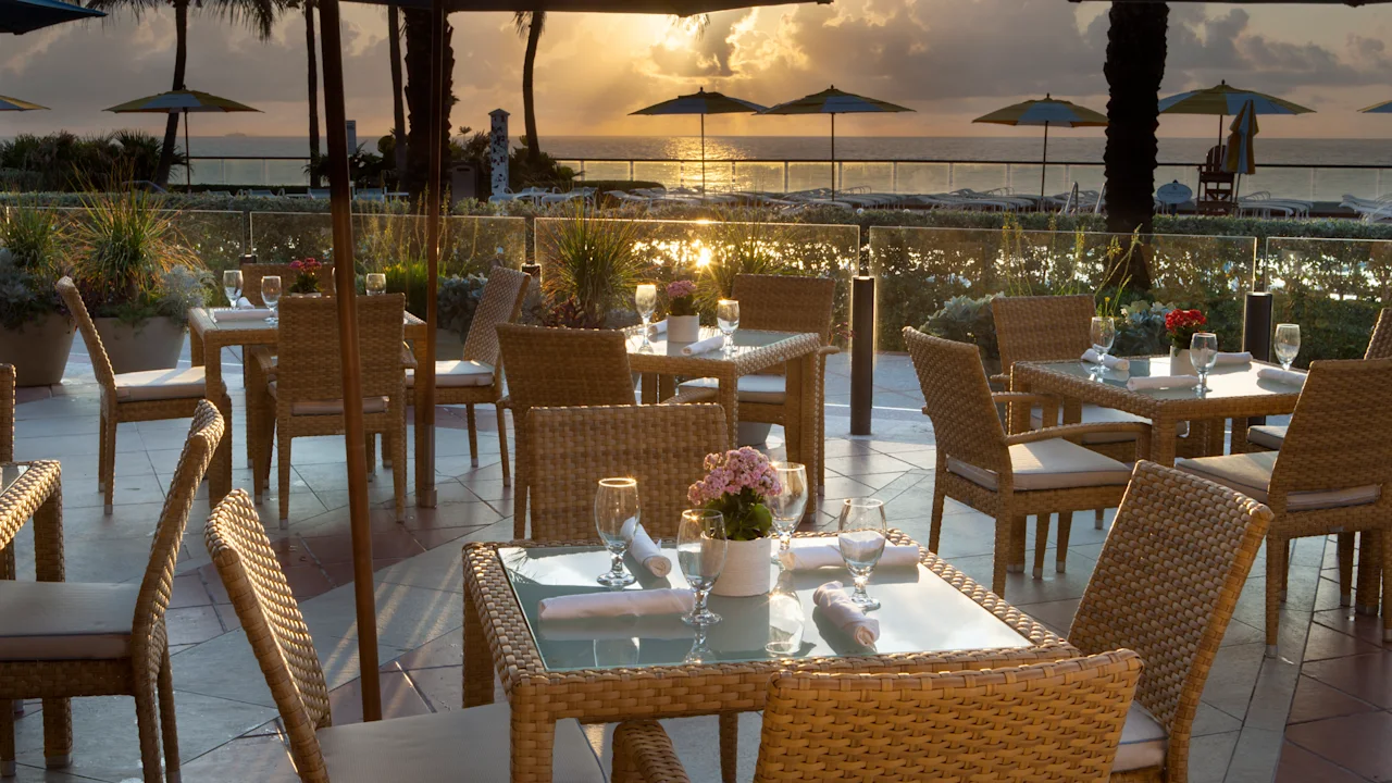 Oceanfront restaurant patio with wicker dining tables at sunset, blue umbrellas and palm trees visible.