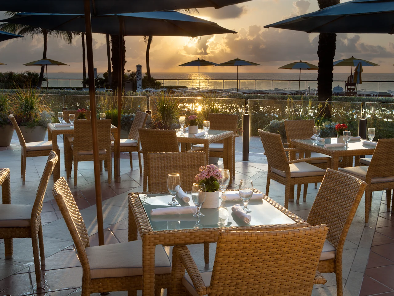 Oceanfront restaurant patio with wicker dining tables at sunset, blue umbrellas and palm trees visible.
