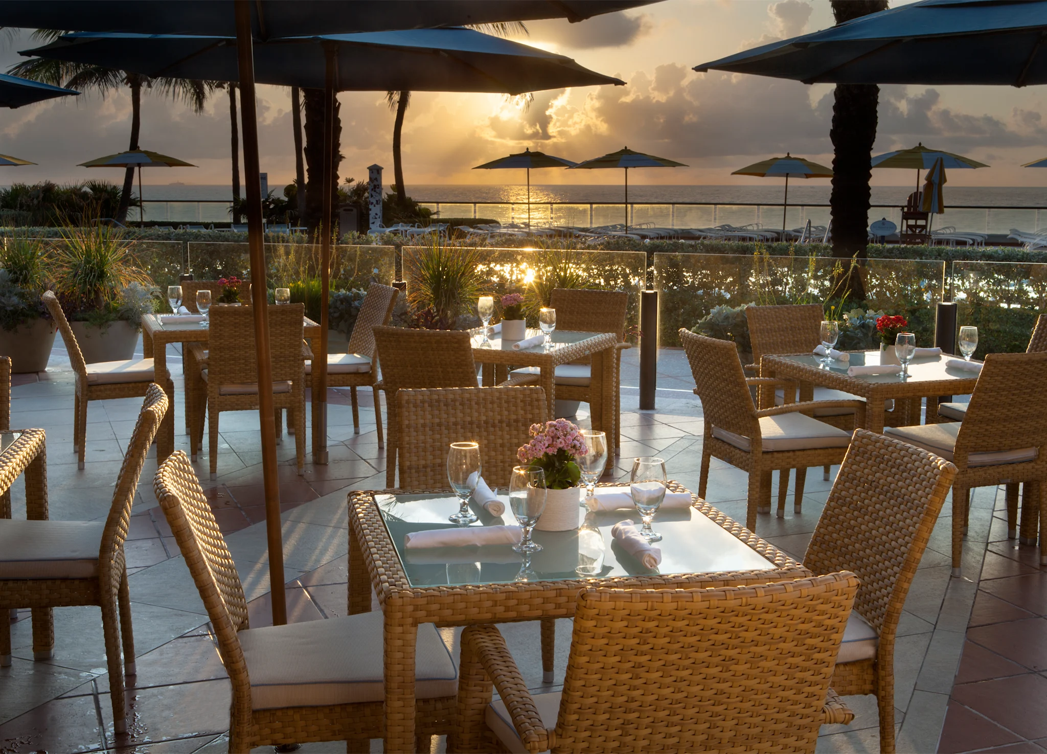 Oceanfront restaurant patio with wicker dining tables at sunset, blue umbrellas and palm trees visible.