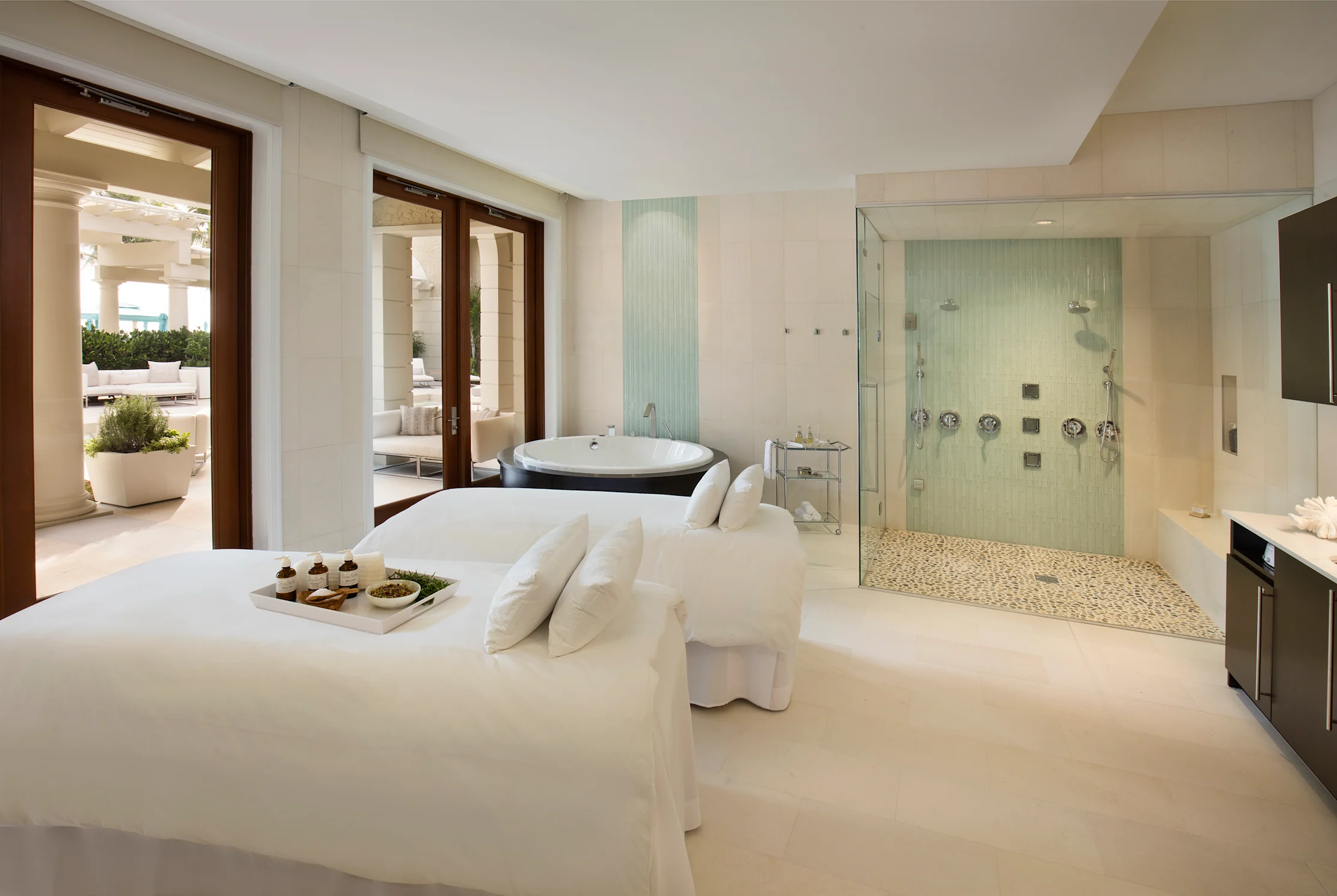 Luxury spa room with white massage bed, glass-enclosed shower, soaking tub, and patio access.