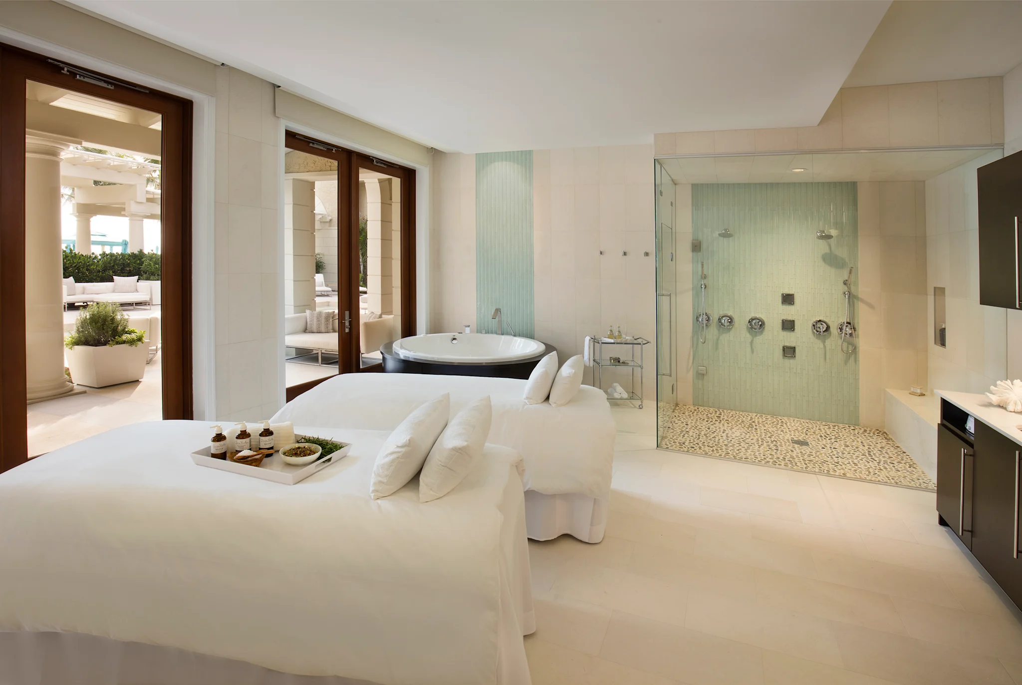 Luxury spa room with white massage bed, glass-enclosed shower, soaking tub, and patio access.