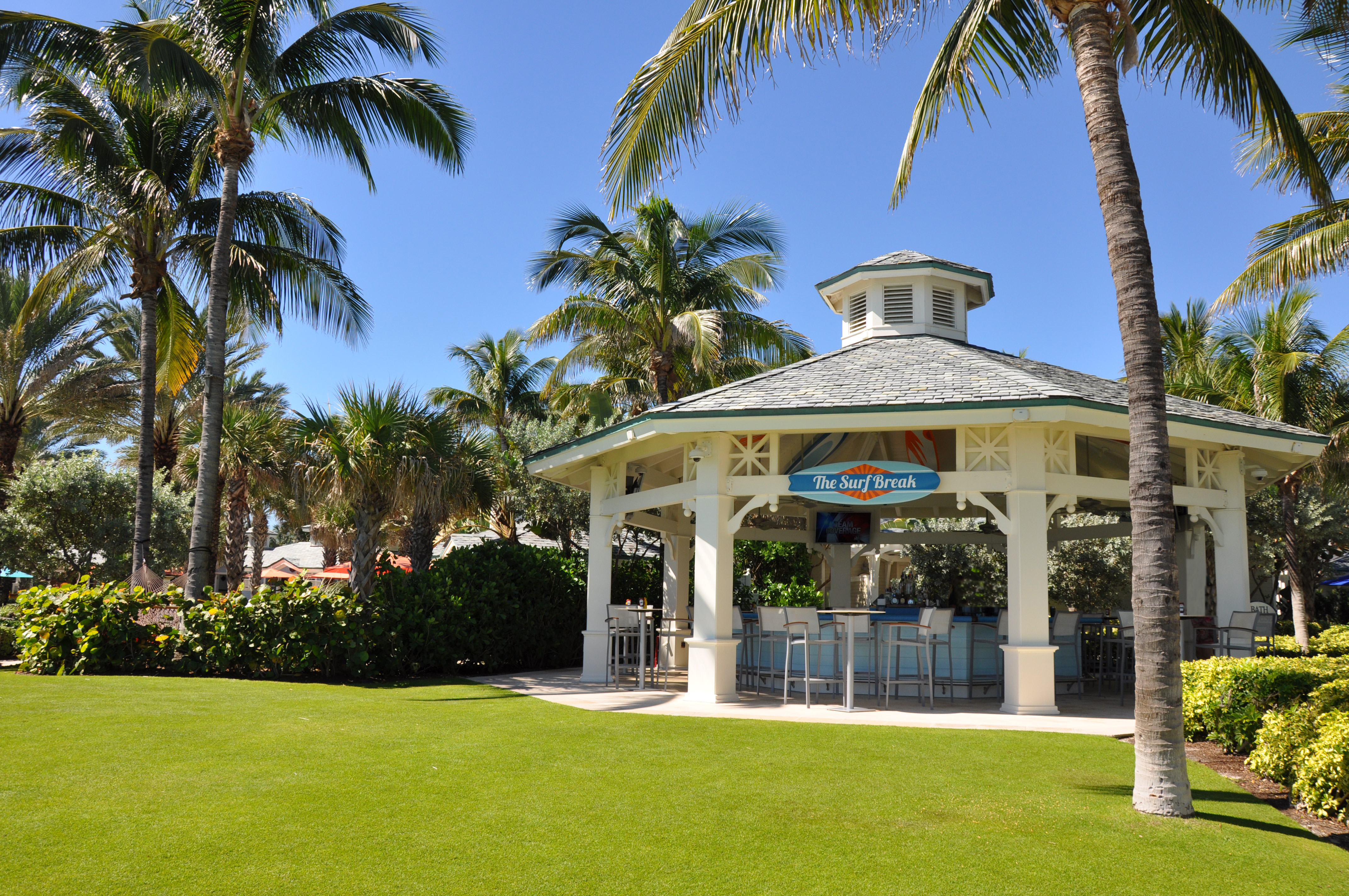 Open-air beach bar pavilion with green roof surrounded by palm trees and manicured lawn under blue sky.