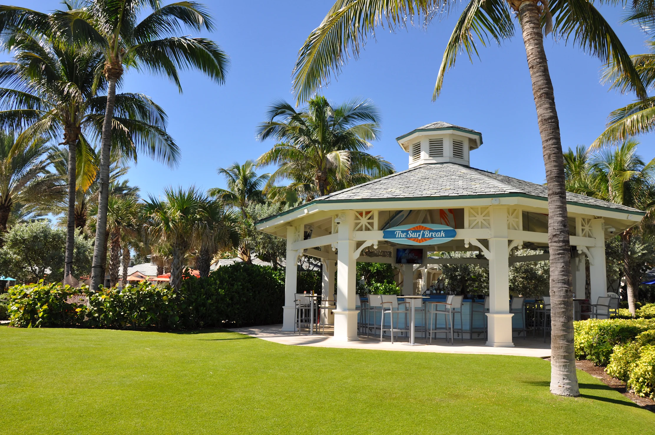 Open-air beach bar pavilion with green roof surrounded by palm trees and manicured lawn under blue sky.