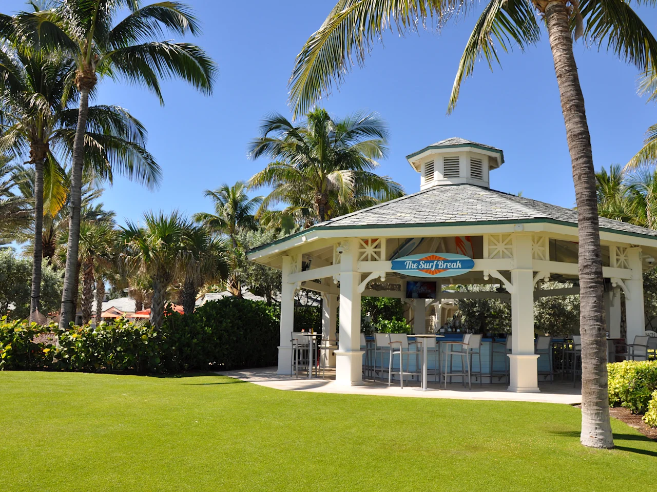 Open-air beach bar pavilion with green roof surrounded by palm trees and manicured lawn under blue sky.