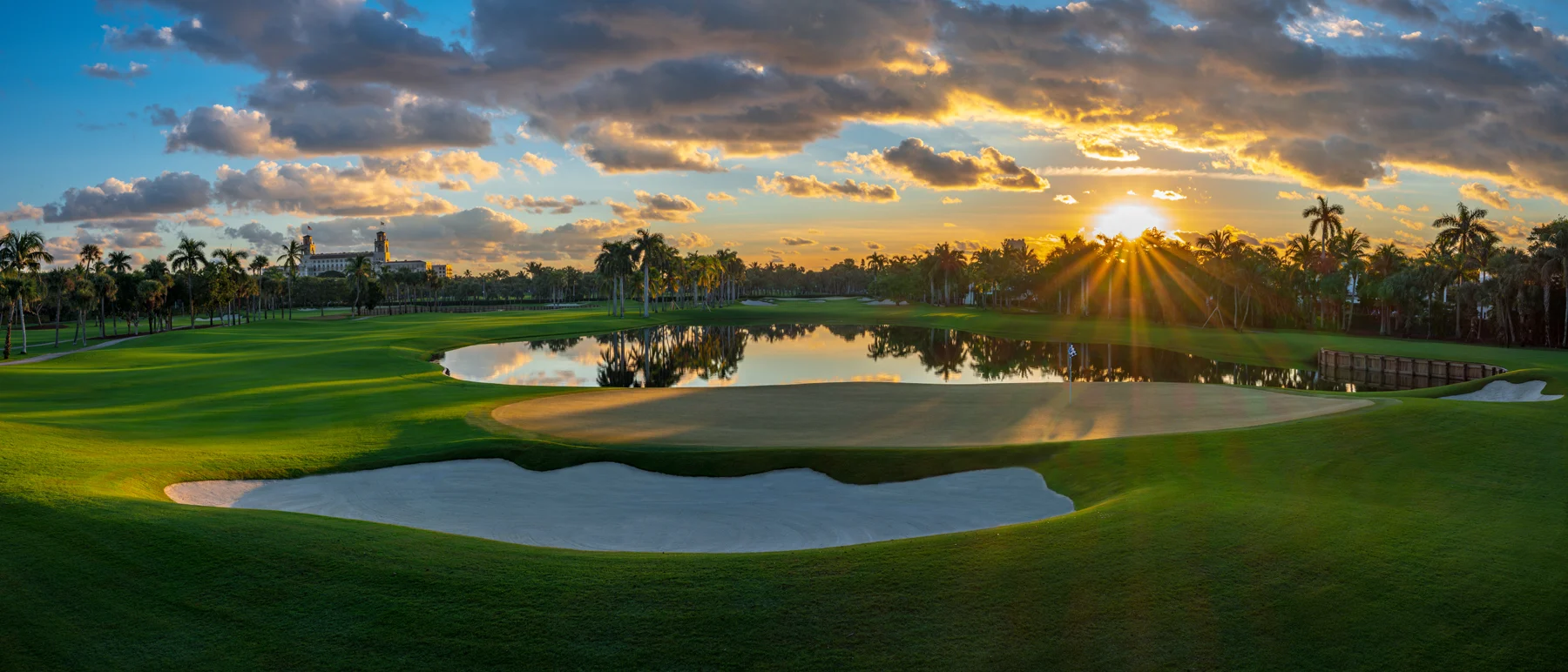 Golf course at sunset with palm trees, water hazards, sand bunkers, and dramatic cloudy sky.