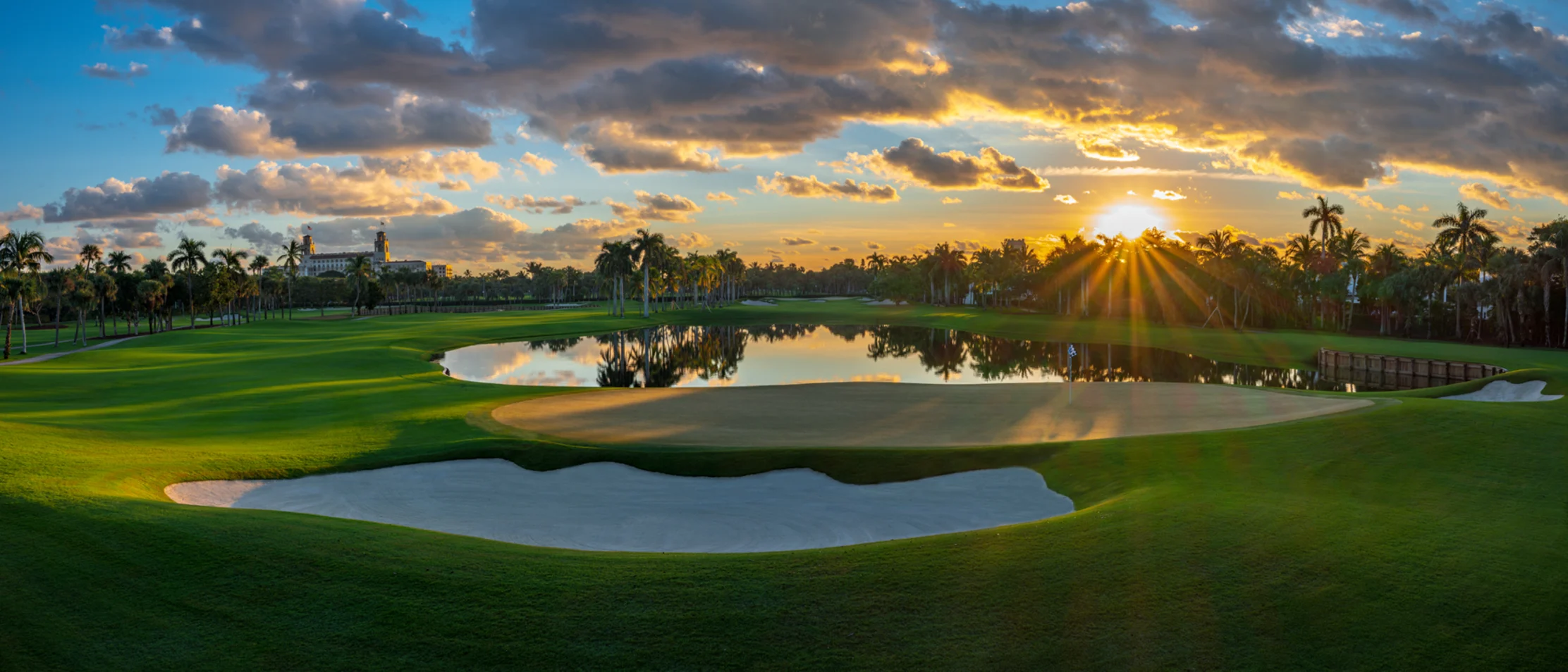 Golf course at sunset with palm trees, water hazards, sand bunkers, and dramatic cloudy sky.