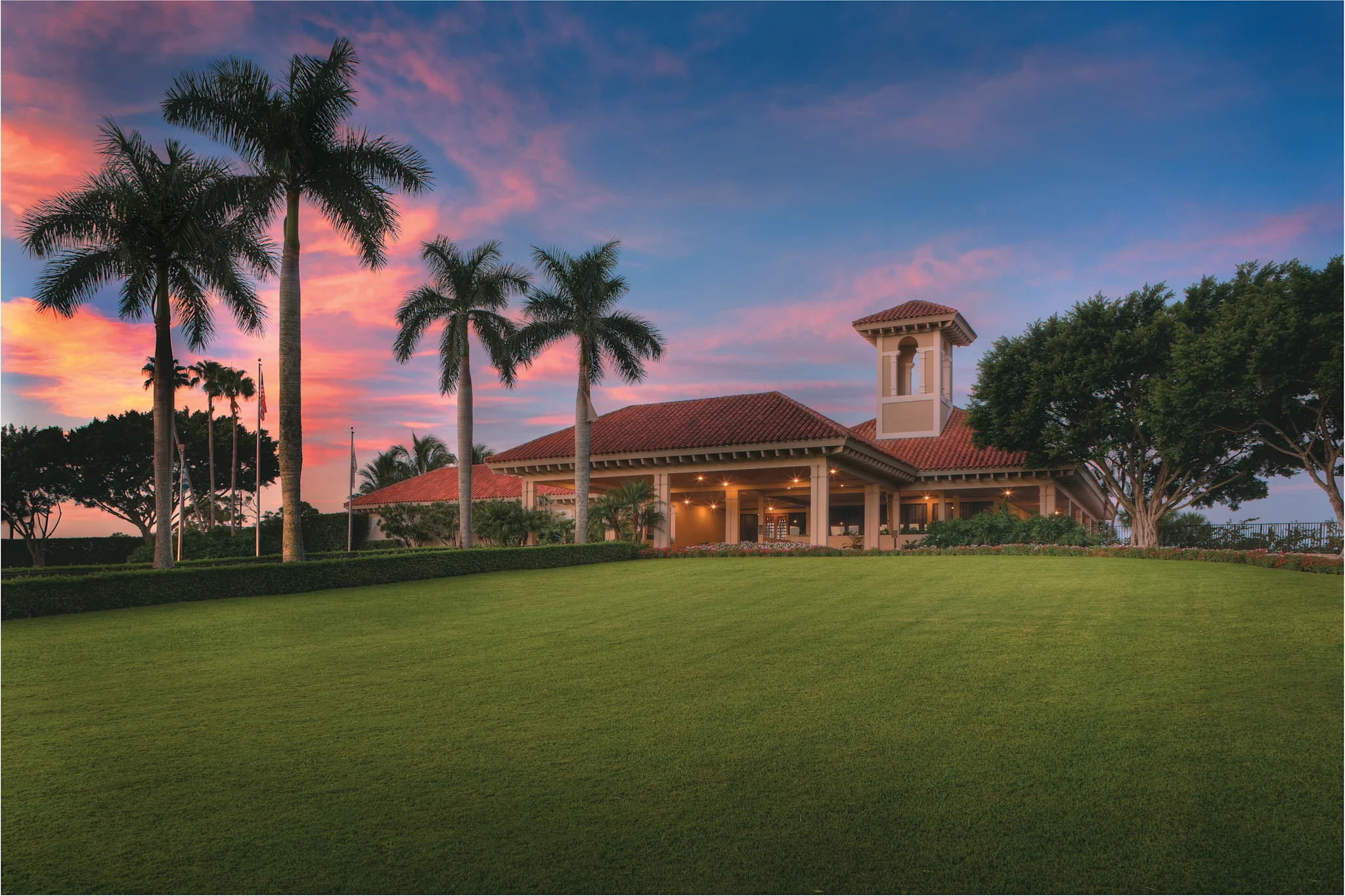 Mediterranean-style clubhouse with red tile roof and bell tower, palm trees, and green lawn at sunset.