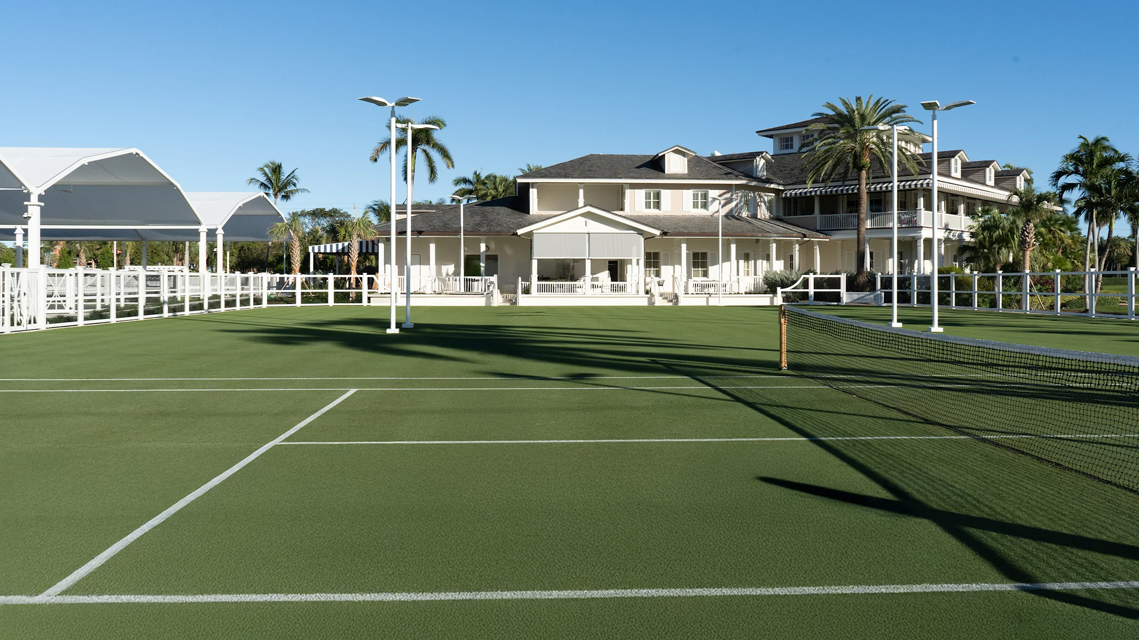 Green tennis courts with white pavilion and beige residential buildings surrounded by palm trees.