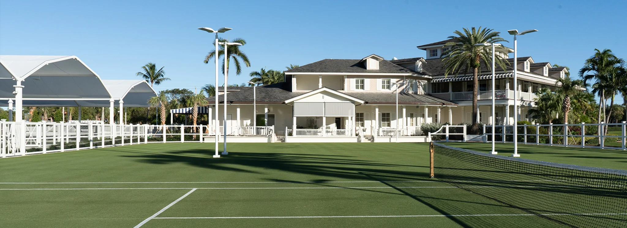 Green tennis courts with white pavilion and beige residential buildings surrounded by palm trees.