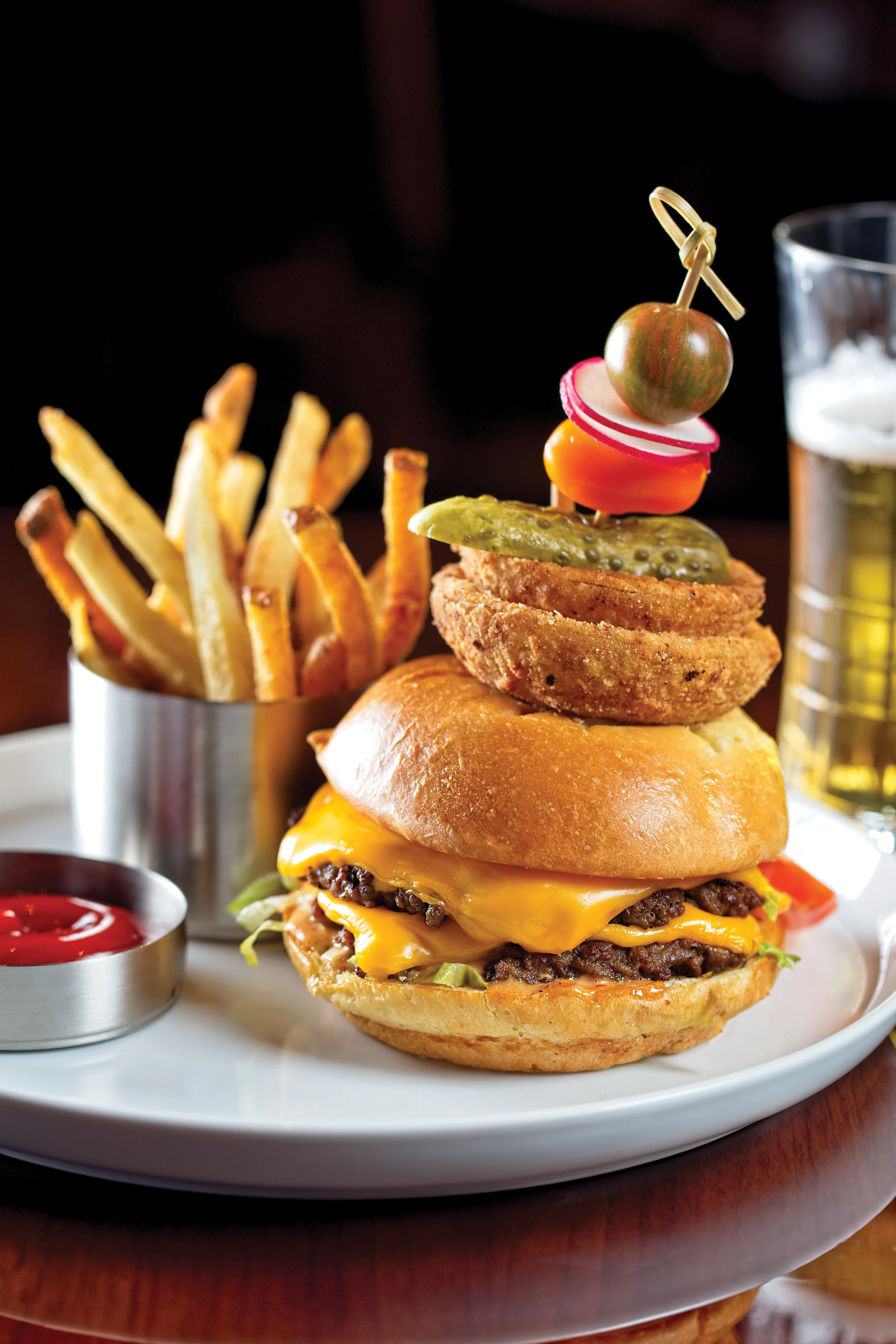 Cheeseburger with onion rings and garnish skewer, served with fries, ketchup, and beer on a plate.