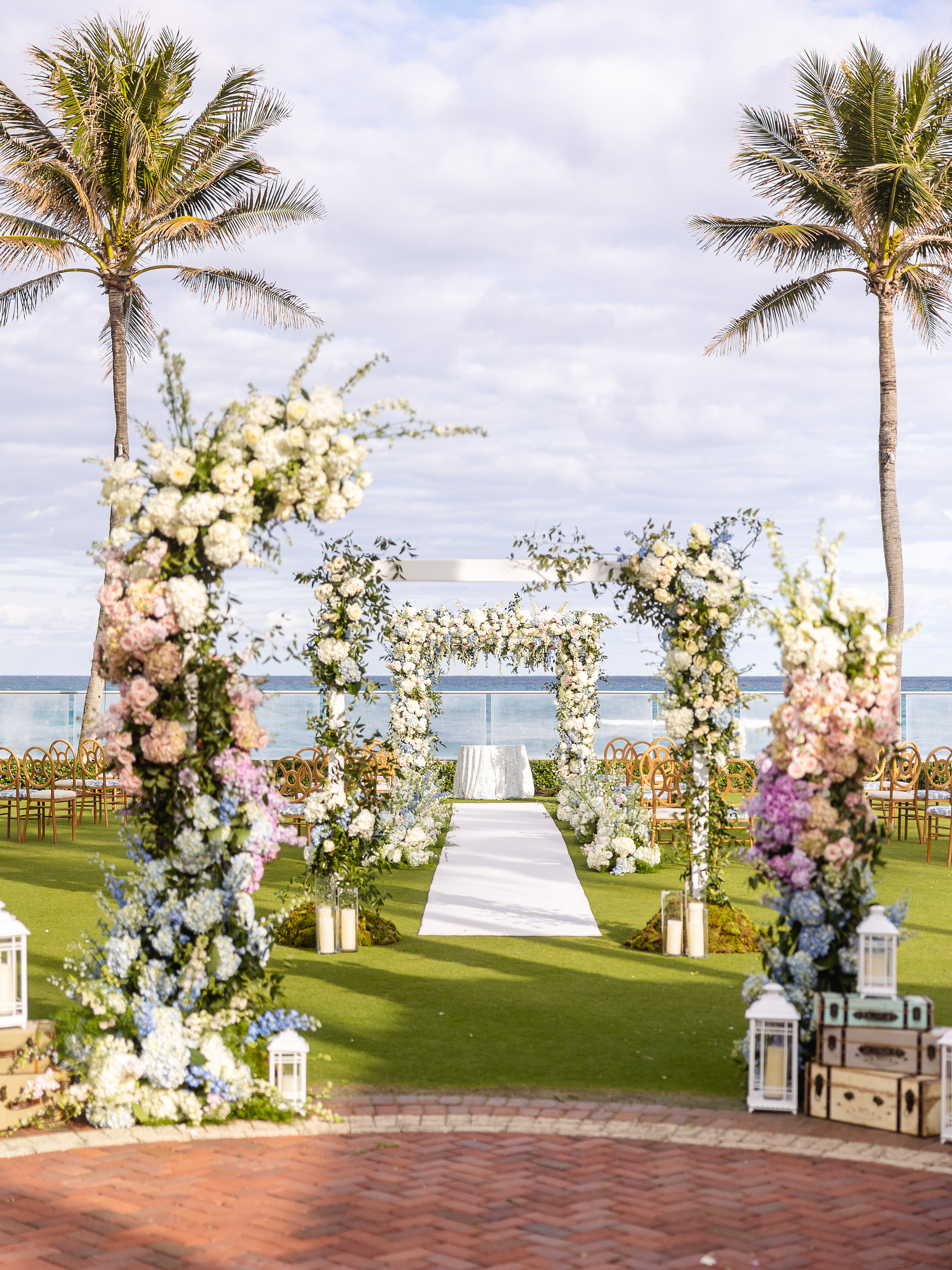 Beachside wedding ceremony setup with floral arch, white aisle runner, palm trees, and ocean view.