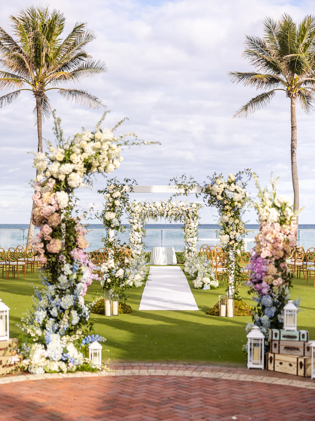Beachside wedding ceremony setup with floral arch, white aisle runner, palm trees, and ocean view.