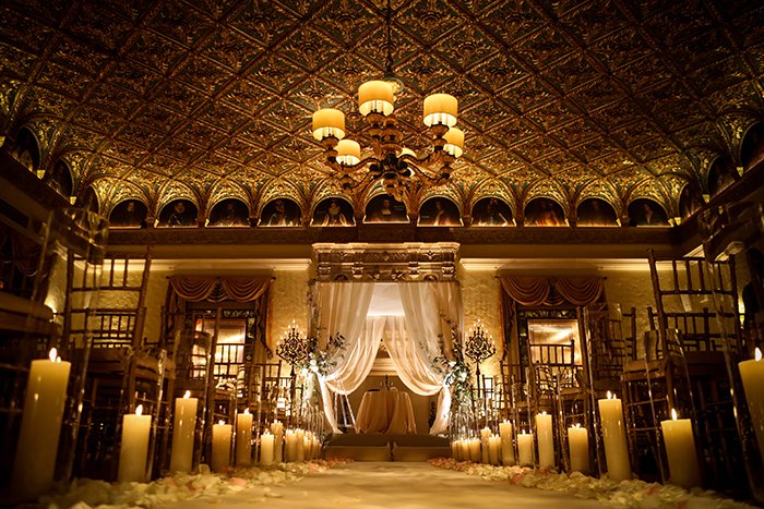 Ornate ballroom with golden ceiling, chandelier, white draped altar, and candles lining the aisle.