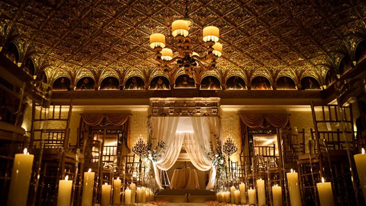 Ornate ballroom with golden ceiling, chandelier, white draped altar, and candles lining the aisle.