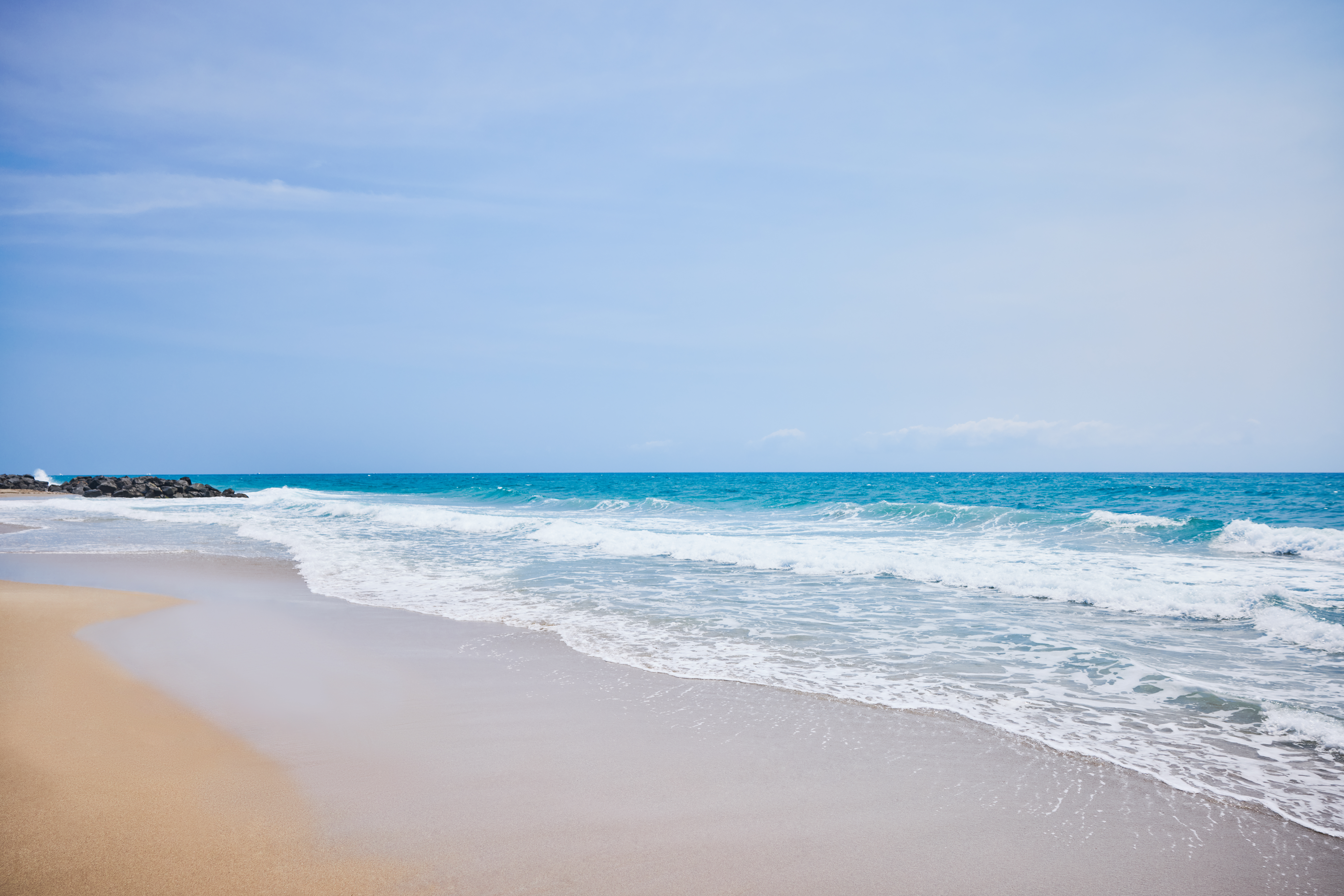 Turquoise ocean waves rolling onto sandy beach with rocks under clear blue sky