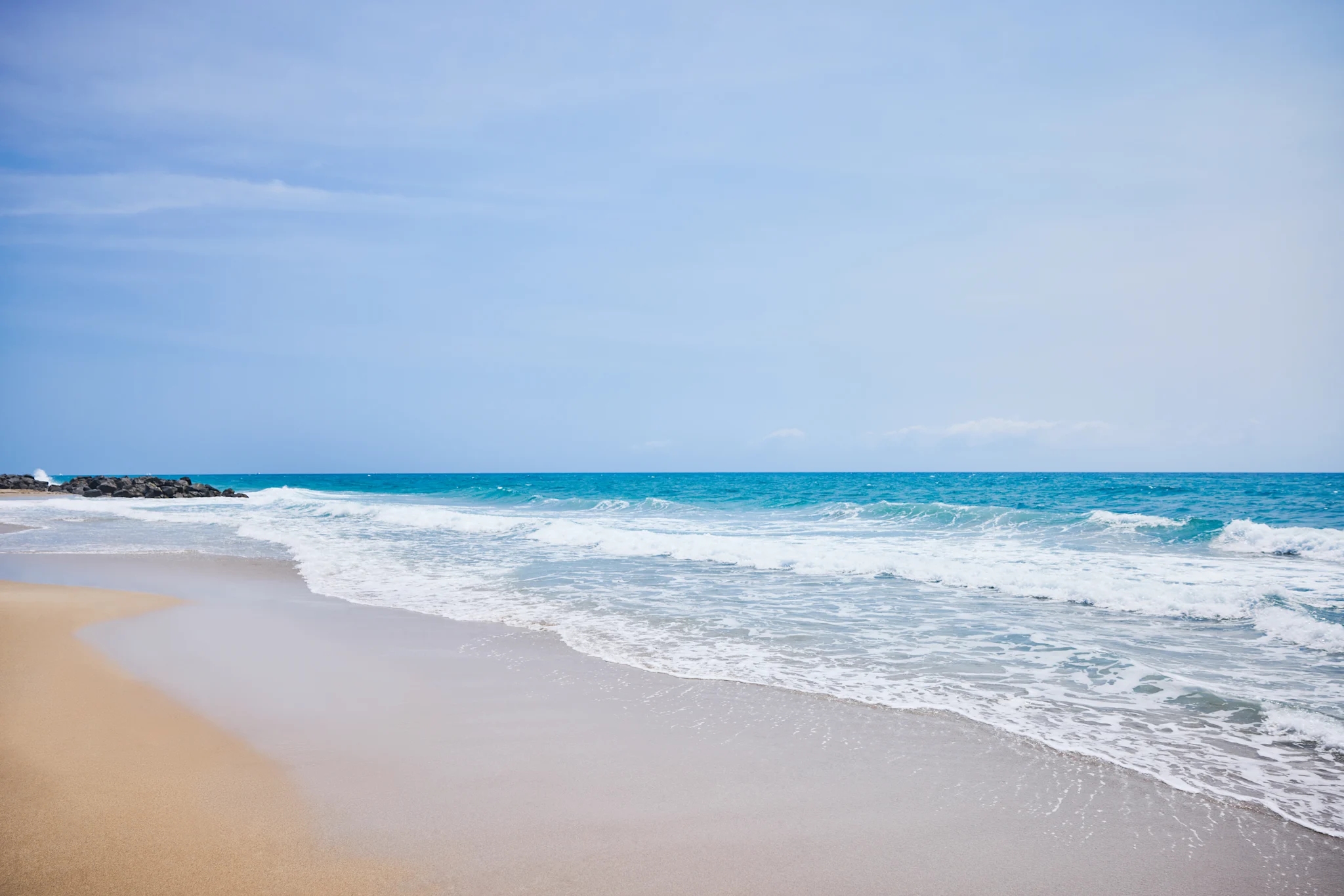 Turquoise ocean waves rolling onto sandy beach with rocks under clear blue sky