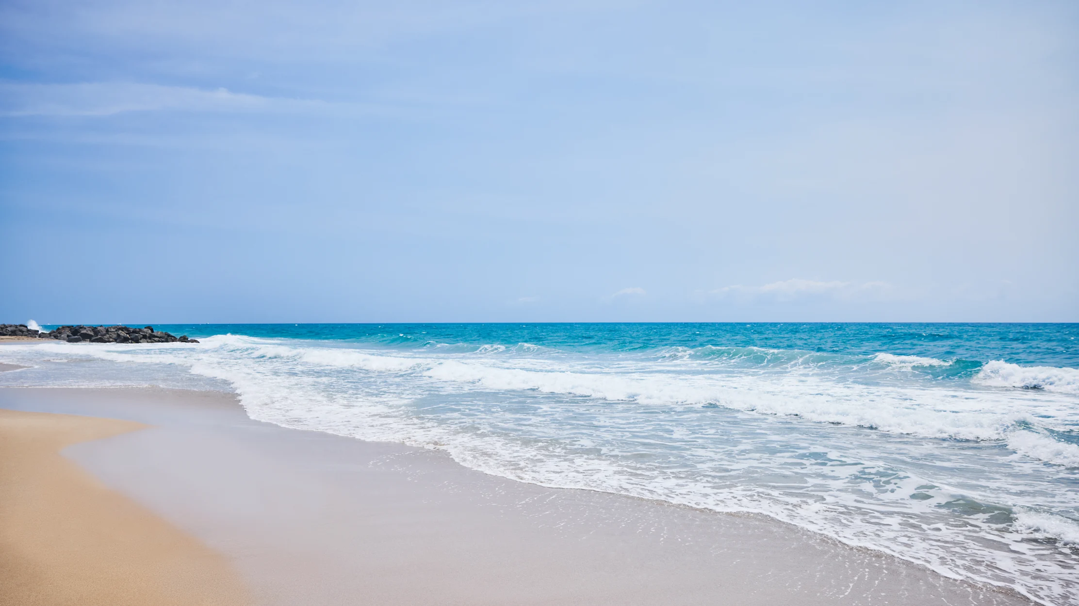Turquoise ocean waves rolling onto sandy beach with rocks under clear blue sky