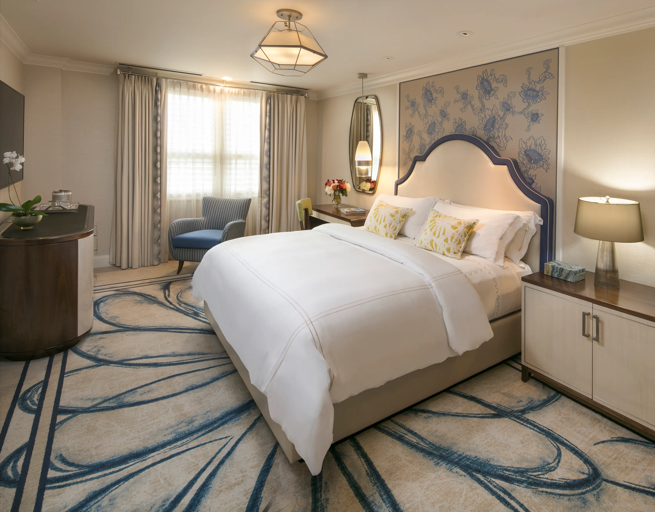 Elegant hotel bedroom with white bedding, blue accent chair, floral wallpaper, and patterned carpet.