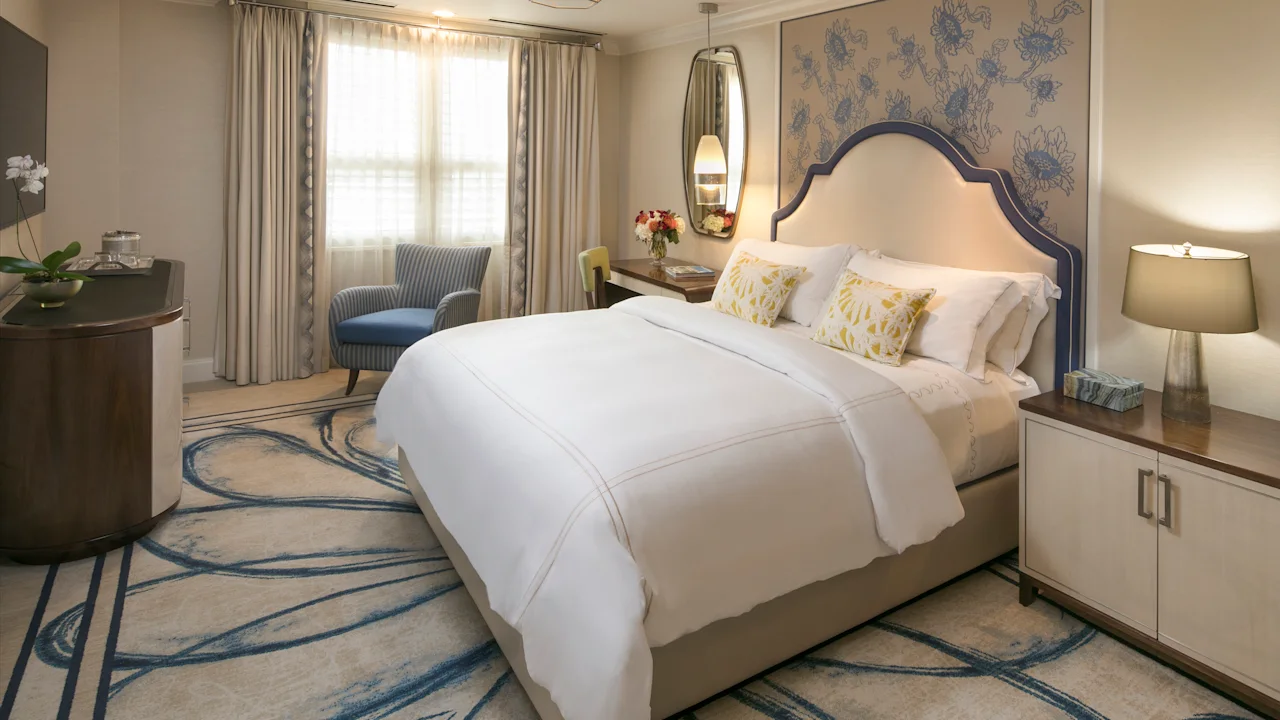 Elegant hotel bedroom with white bedding, blue accent chair, floral wallpaper, and patterned carpet.