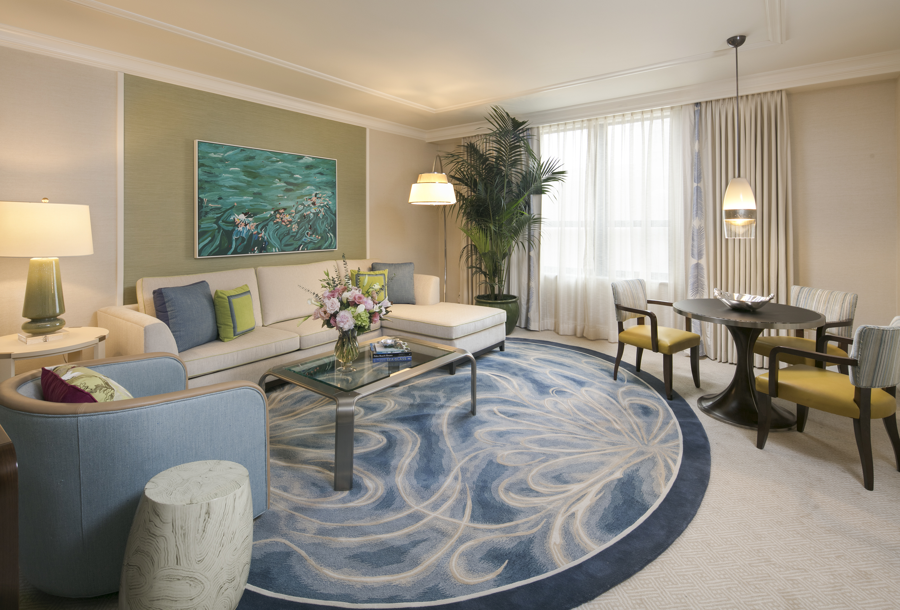 Modern hotel suite with beige and blue decor, ocean artwork, swirl-patterned rug, and dining area.