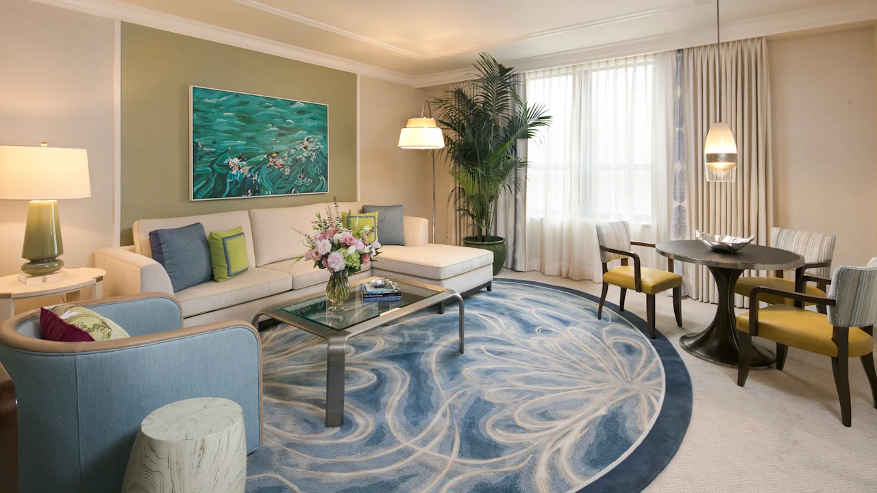Modern hotel suite with beige and blue decor, ocean artwork, swirl-patterned rug, and dining area.