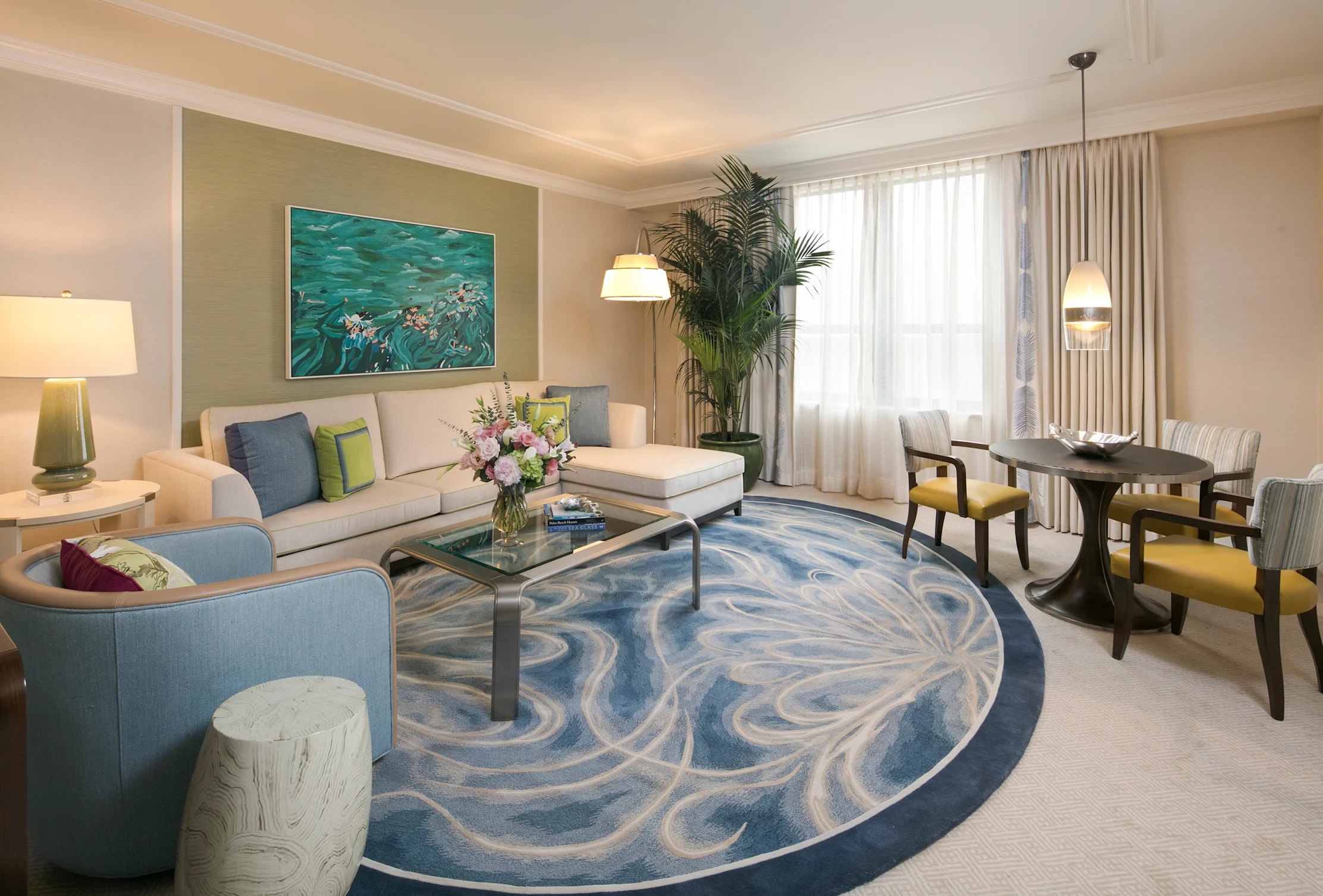 Modern hotel suite with beige and blue decor, ocean artwork, swirl-patterned rug, and dining area.