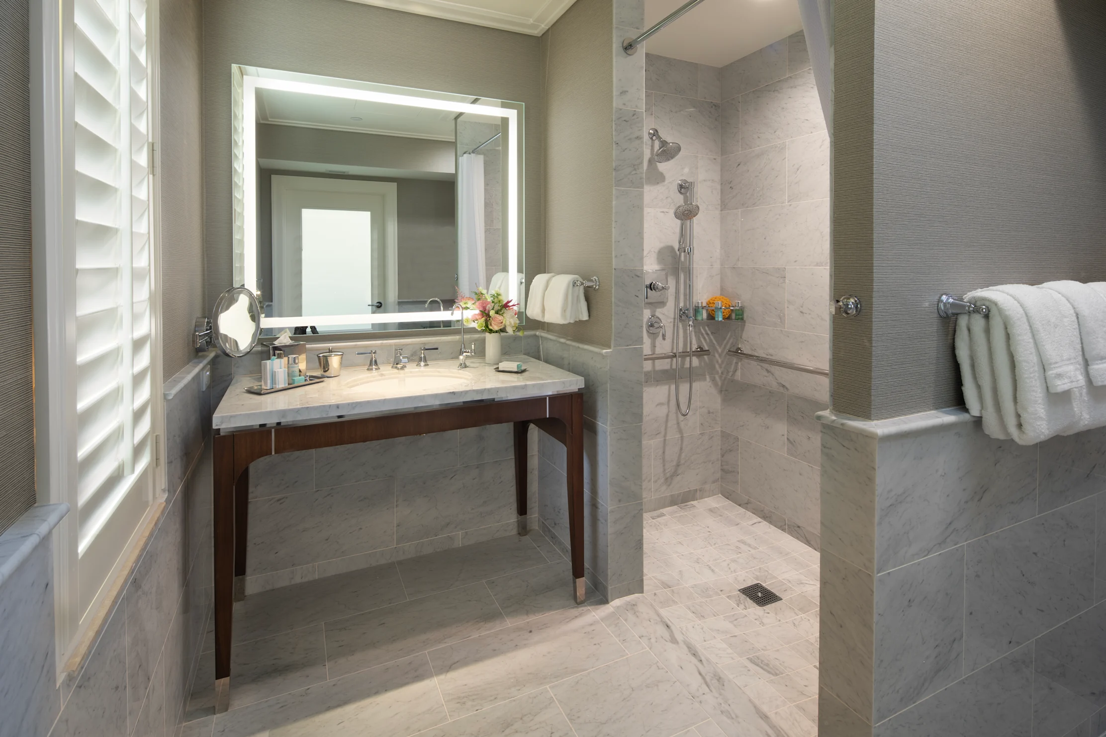 Modern bathroom with gray tile, backlit mirror, marble vanity, walk-in shower, and white towels.