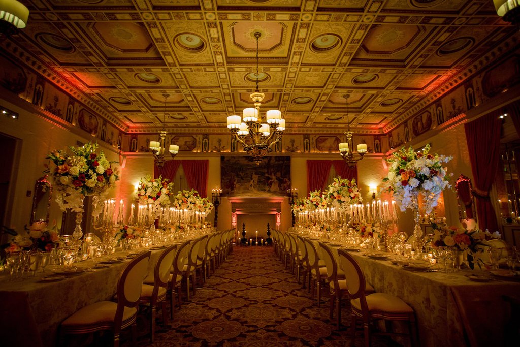 Elegant ballroom with ornate gold ceiling, chandelier, long dining tables with floral centerpieces and candles.