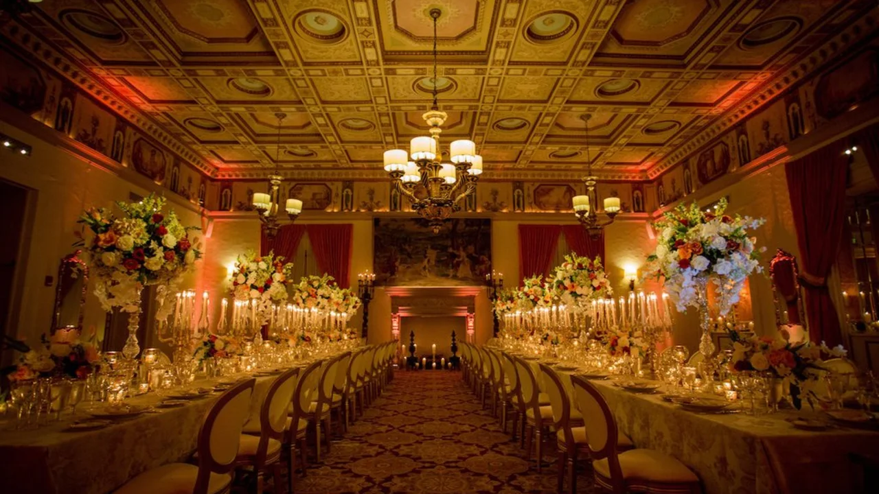 Elegant ballroom with ornate gold ceiling, chandelier, long dining tables with floral centerpieces and candles.