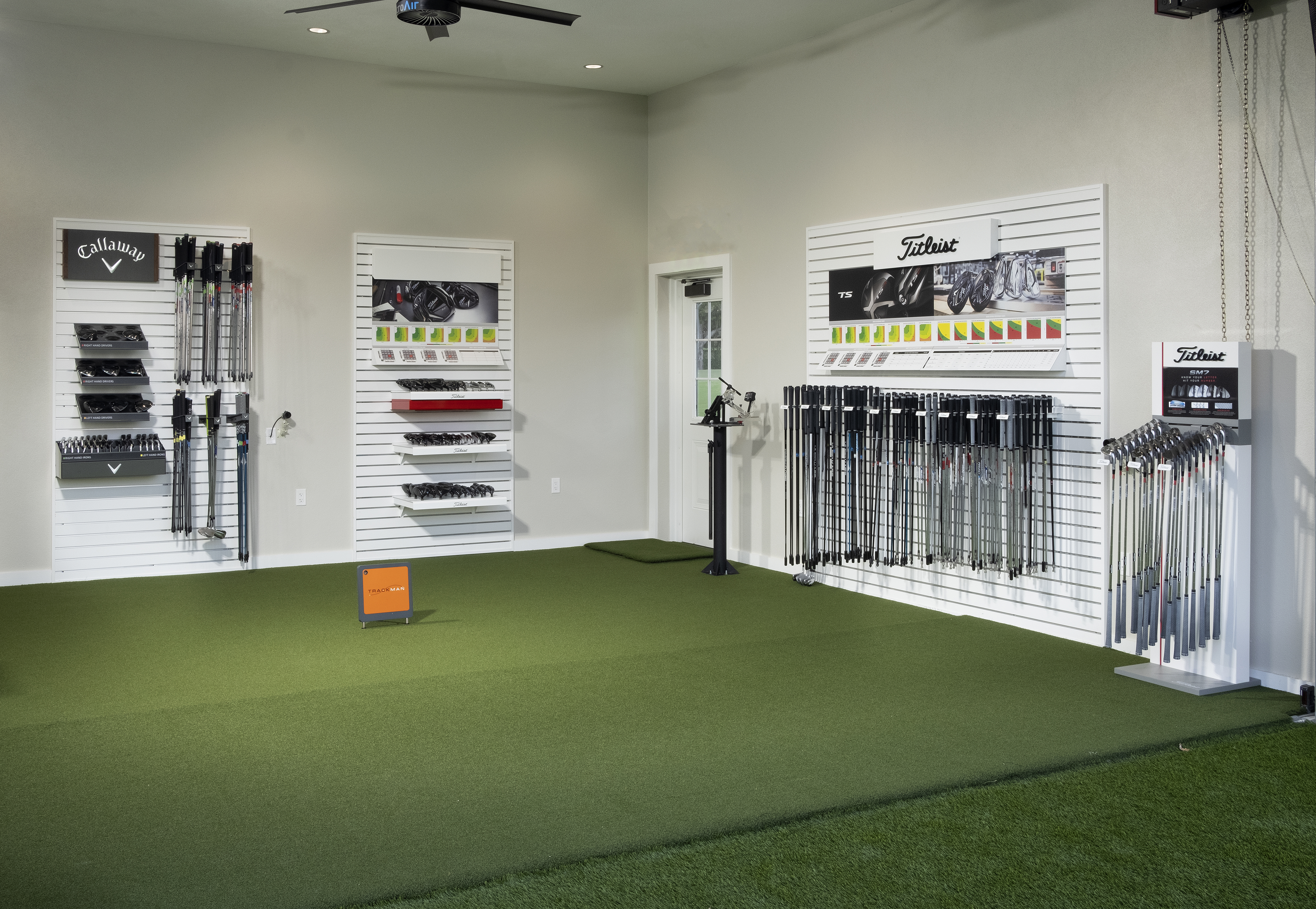 Indoor golf fitting studio with green turf, white slatwall displays of Callaway and Titleist clubs.