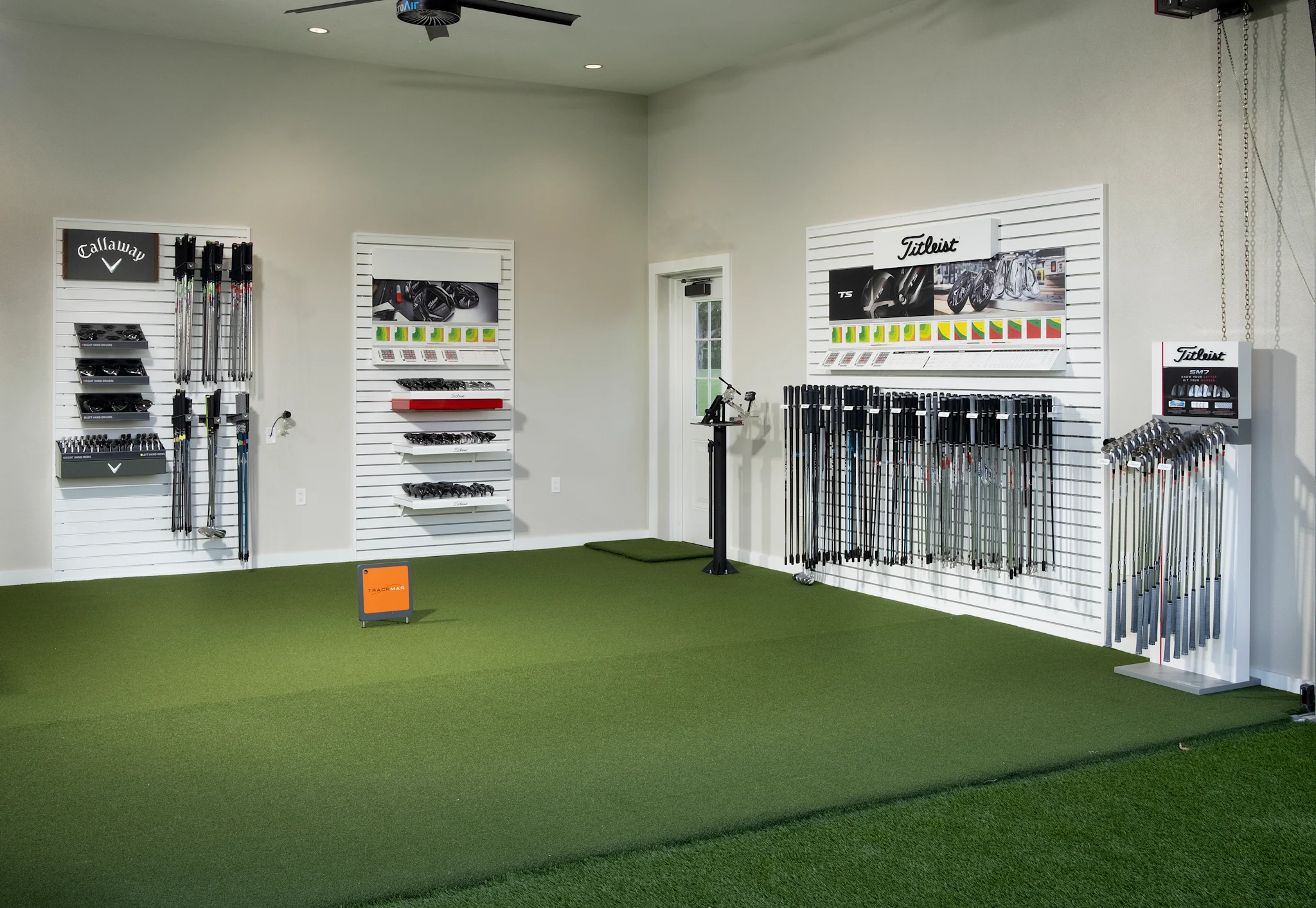 Indoor golf fitting studio with green turf, white slatwall displays of Callaway and Titleist clubs.