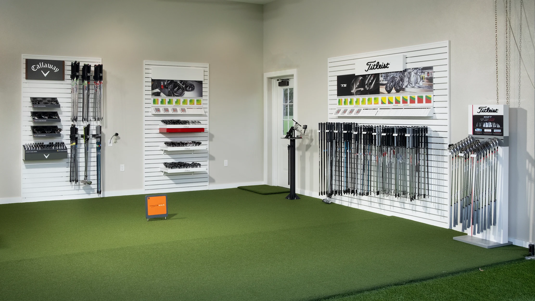 Indoor golf fitting studio with green turf, white slatwall displays of Callaway and Titleist clubs.