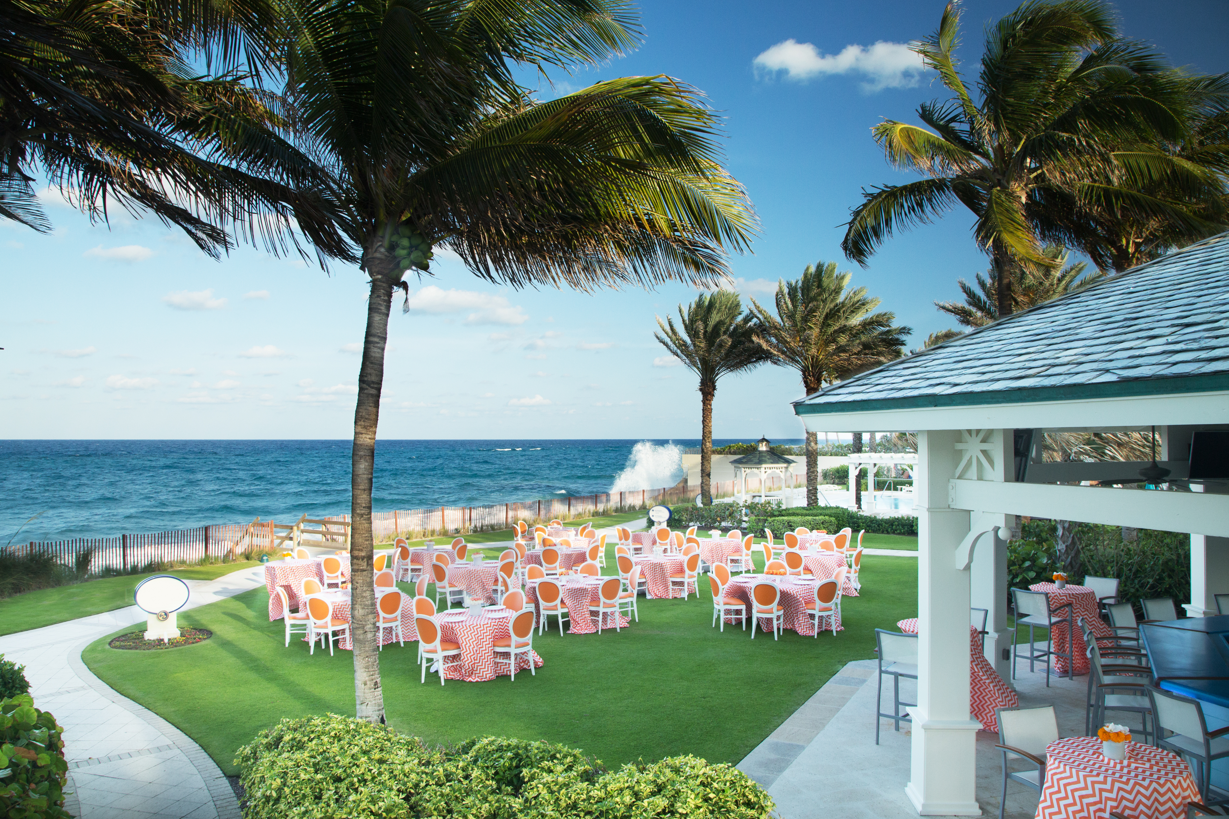 Oceanfront event venue with palm trees, colorful tables on green lawn, and blue ocean waves.