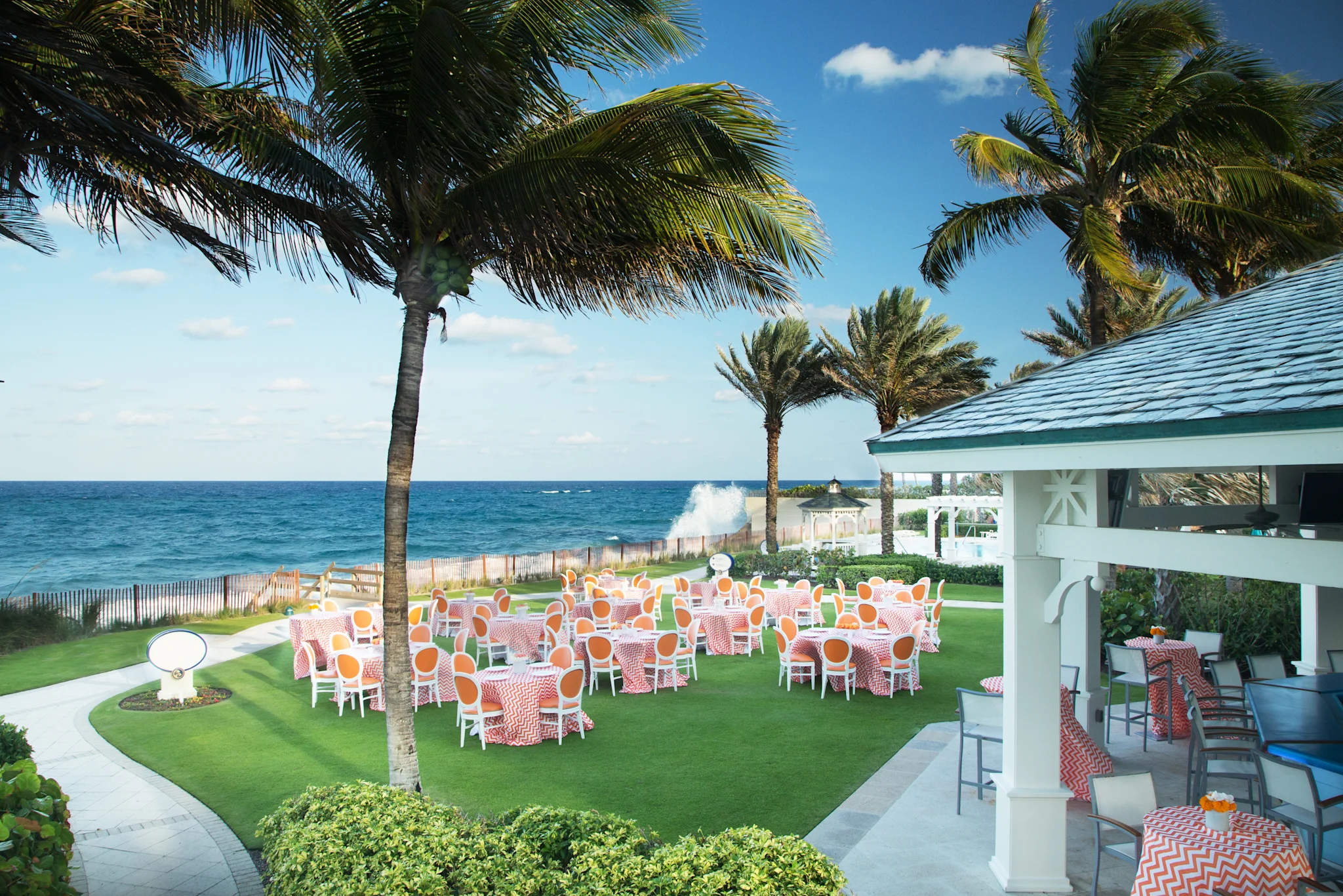 Oceanfront event venue with palm trees, colorful tables on green lawn, and blue ocean waves.