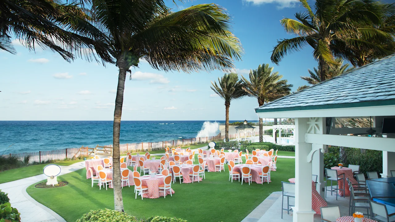 Oceanfront event venue with palm trees, colorful tables on green lawn, and blue ocean waves.
